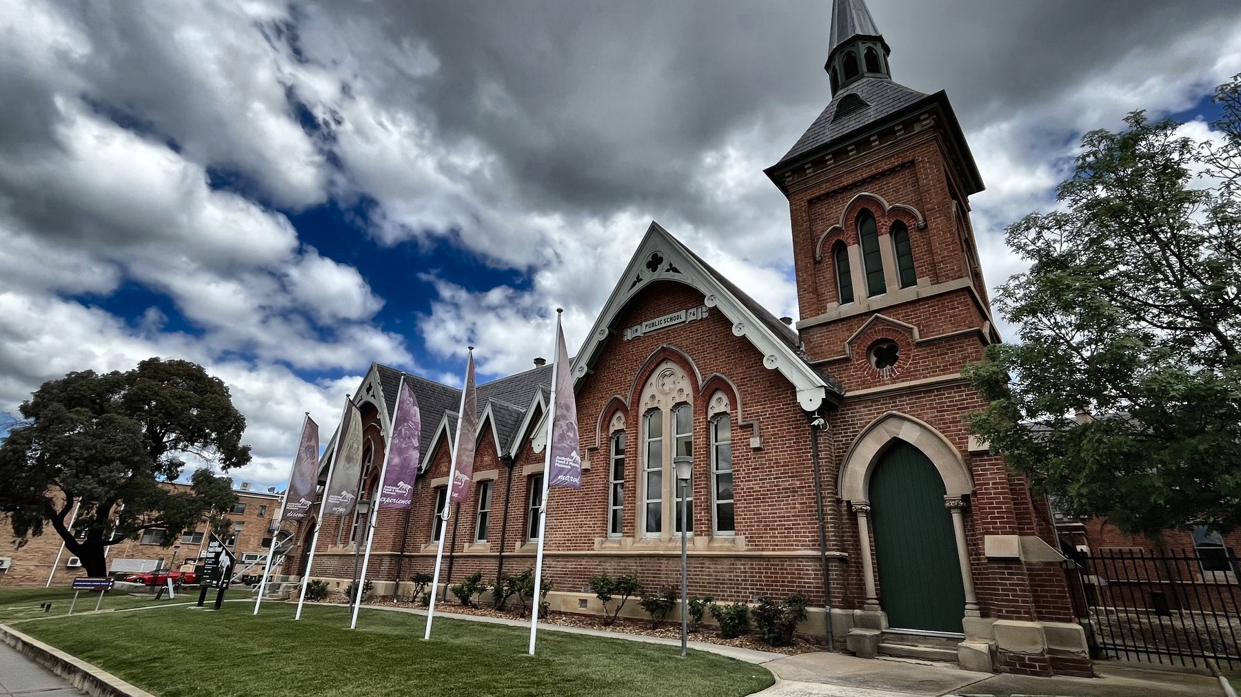A red brick Victorian school building with arched windows, a bell tower and flagpoles along the street frontage