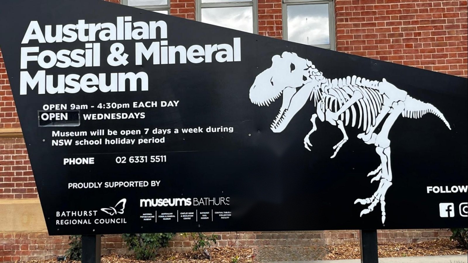 A large black sign outside a brick building reading Australian Fossil and Mineral Museum, open 9am to 4.30pm each day, with a white dinosaur skeleton graphic