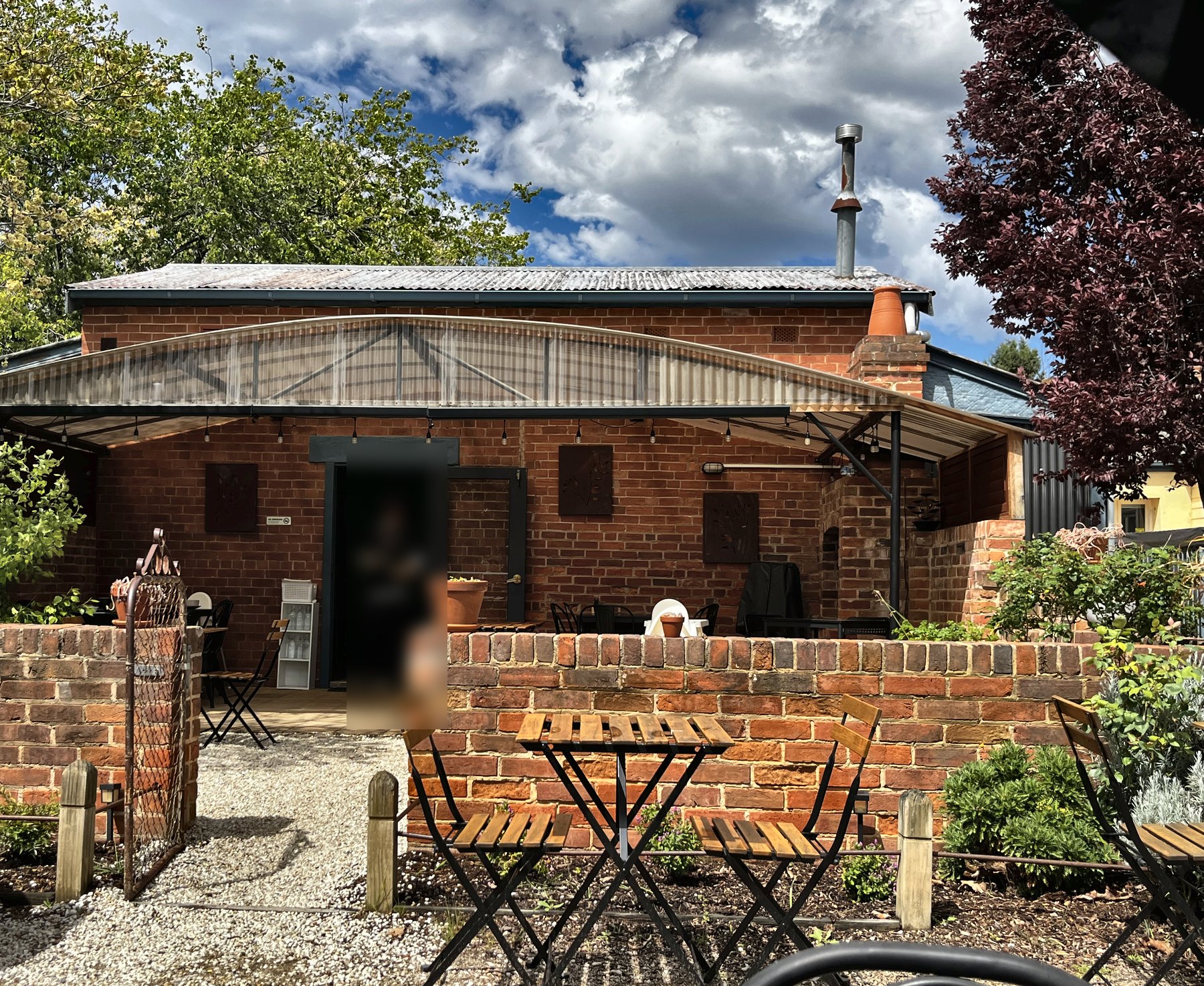 A walled brick courtyard behind the cafe, with timber bistro tables and chairs on gravel under an awning