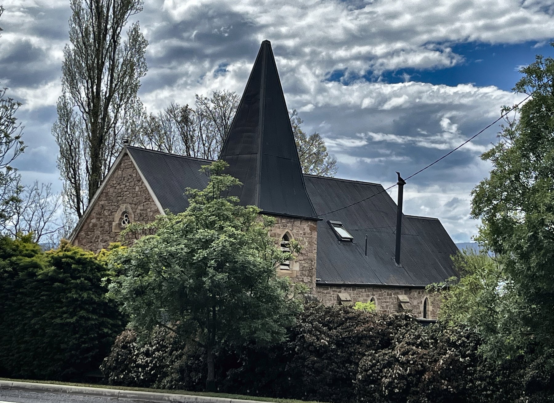 A small stone church with a very steep black shingled spire behind a hedge
