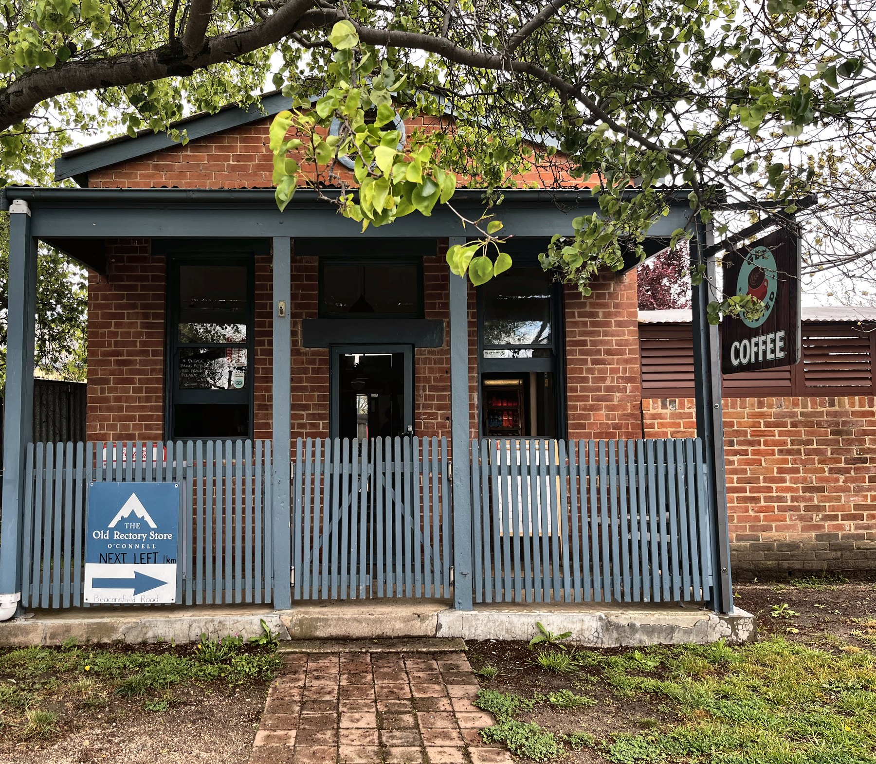 A small brick cafe behind a blue timber picket fence, with a hanging sign reading COFFEE at the side of the building