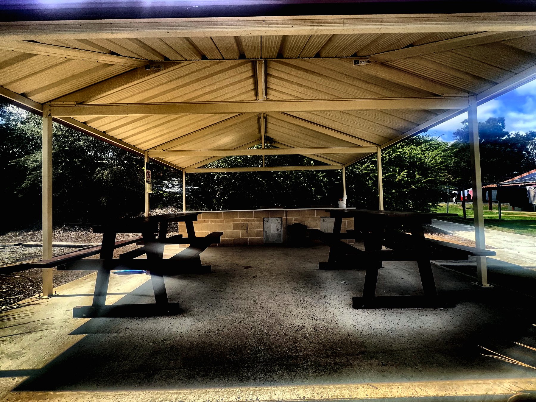 A large open-sided shelter with a pitched roof over two timber picnic tables on concrete