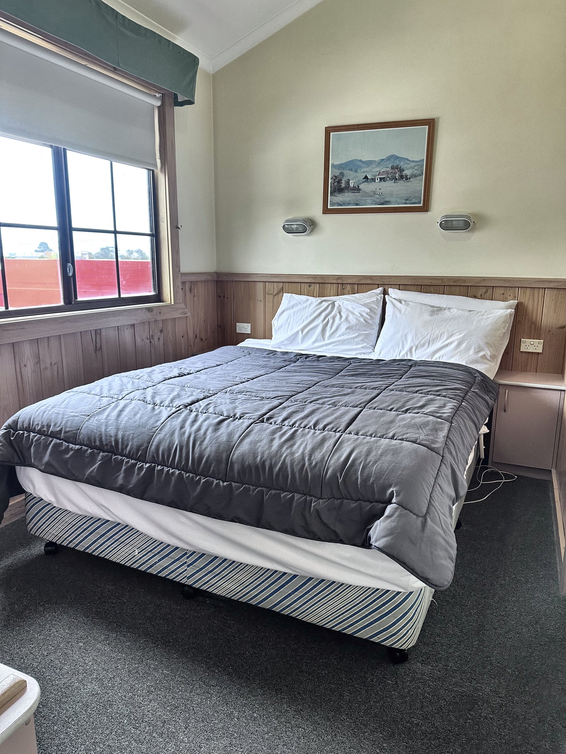 A main bedroom with a queen bed made up with a grey quilt, a framed print and a window