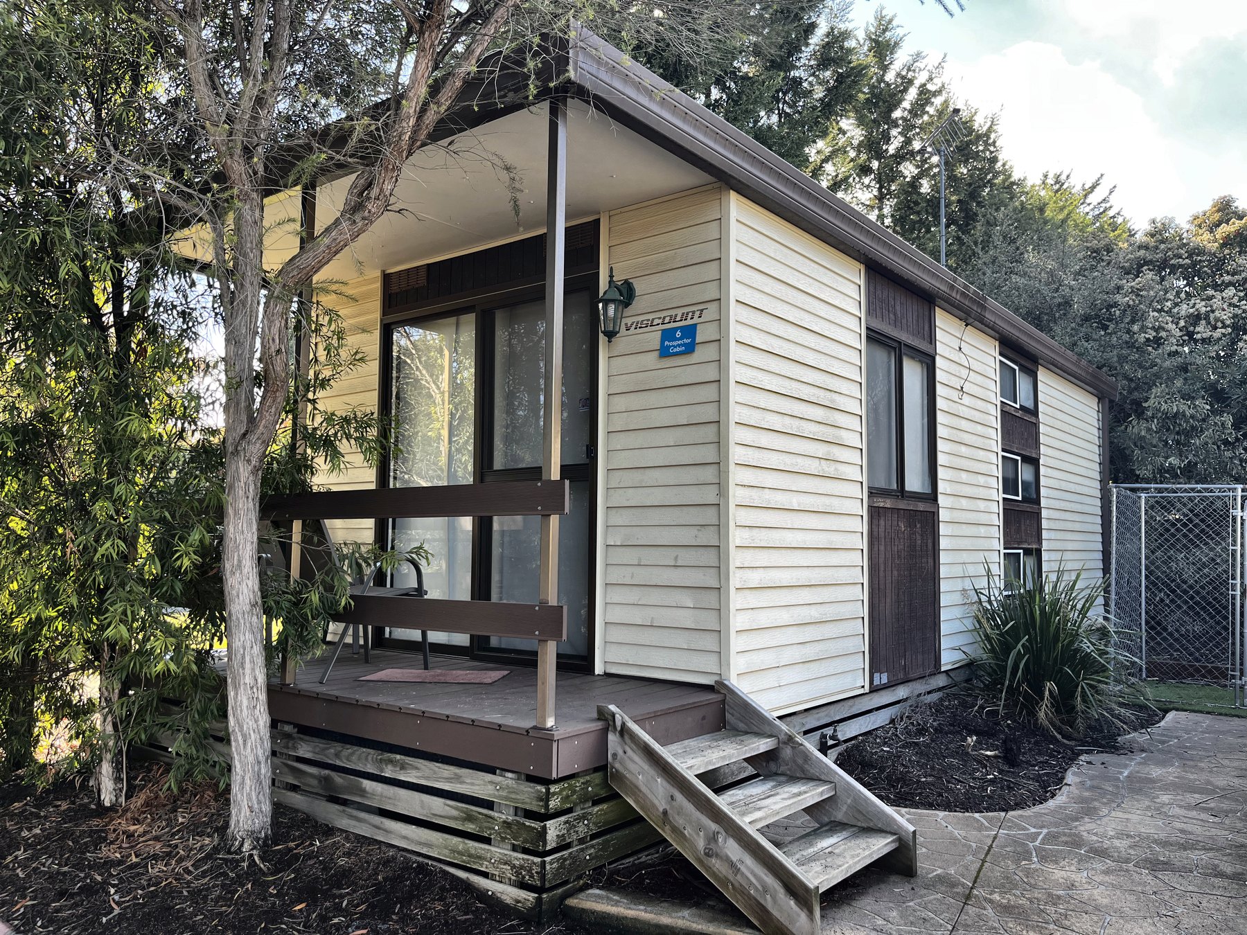 A plainer cream weatherboard cabin with a flat verandah and timber steps under gum trees