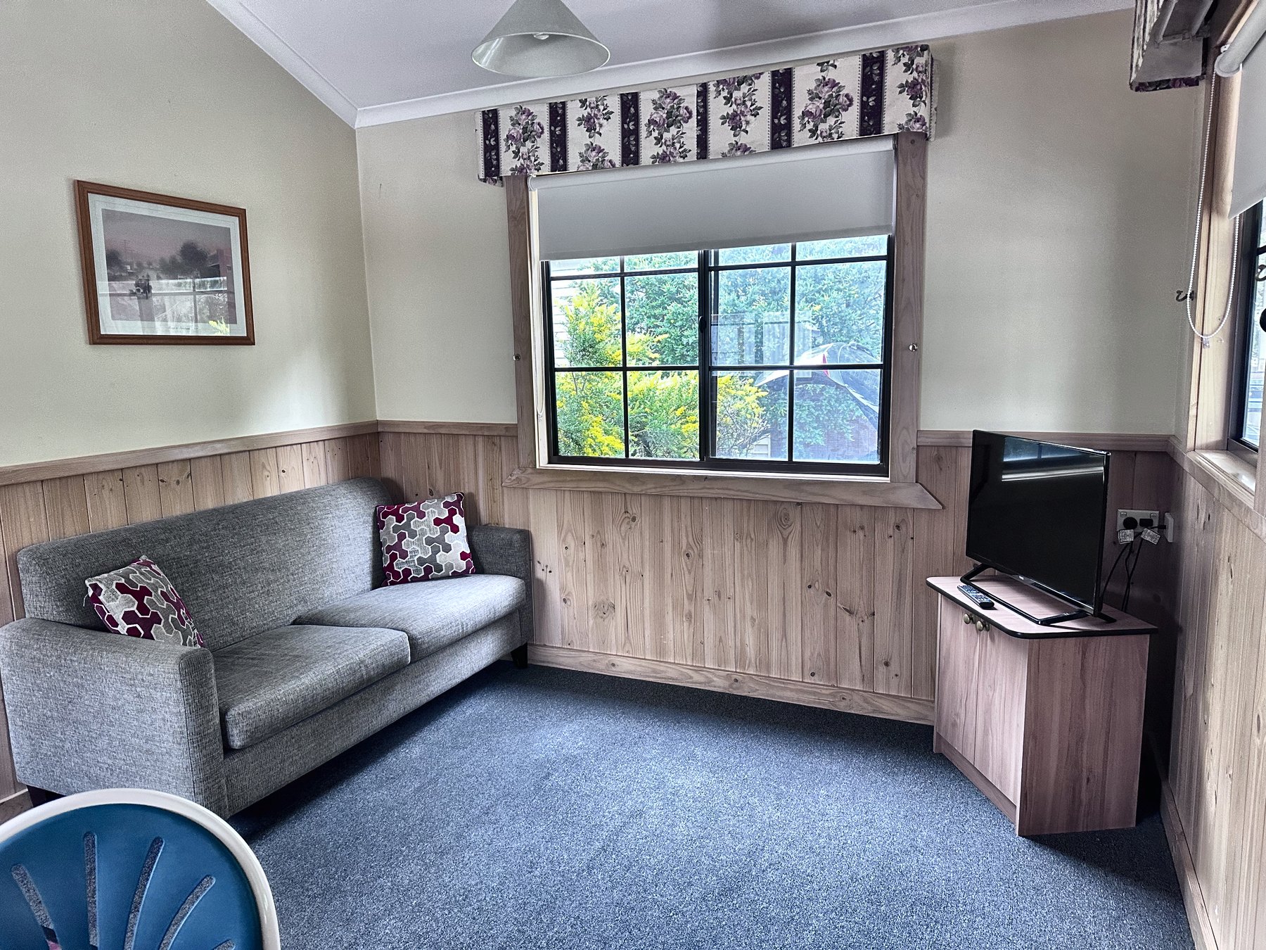 A cabin living room with a grey sofa, a wall-mounted television on a timber unit, carpet and a curtained window