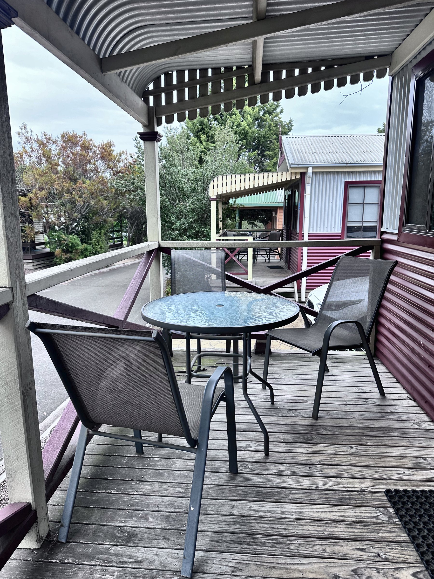 A timber verandah under a corrugated roof with a round glass table and two mesh chairs, looking out over the park