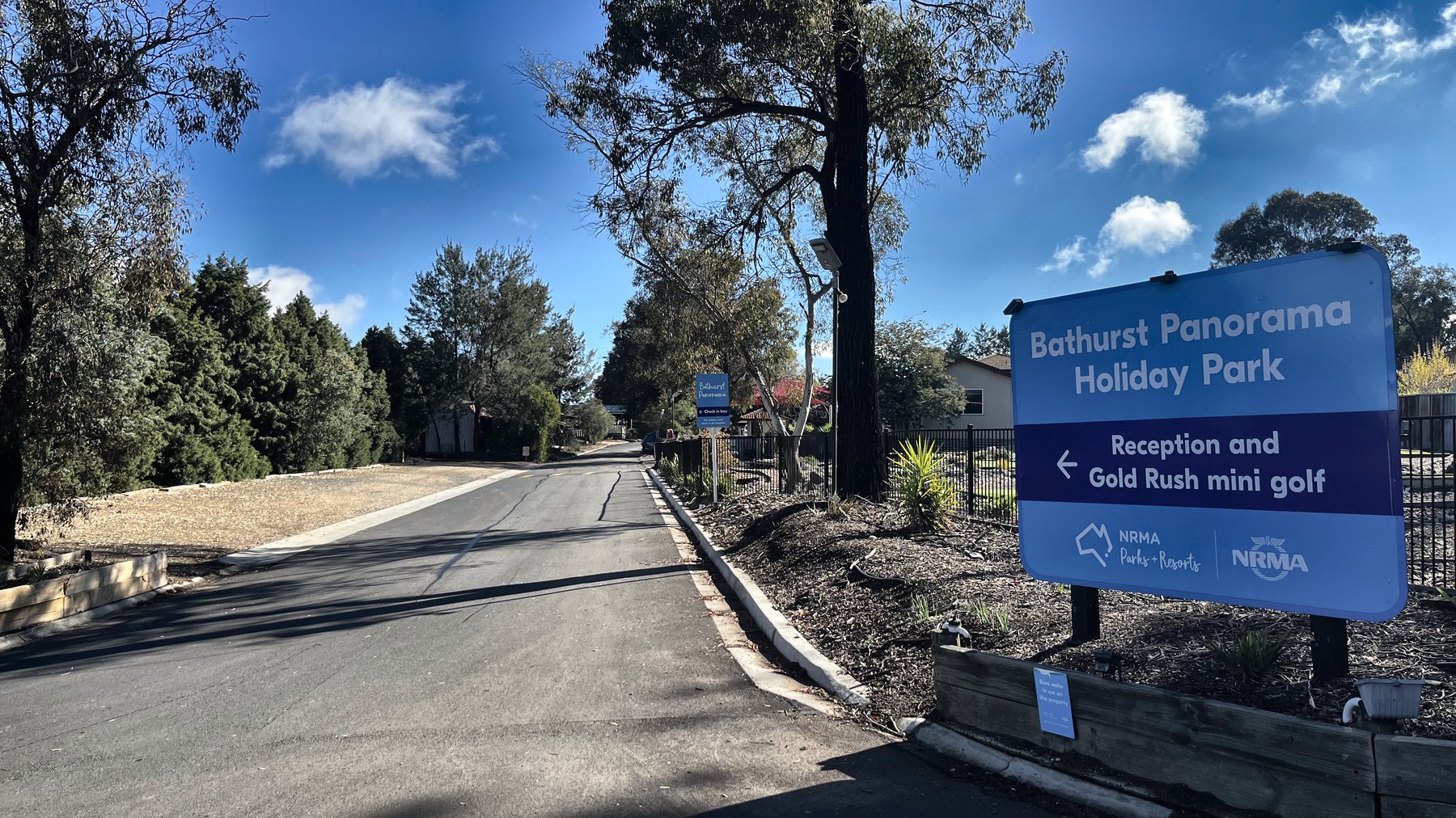 A blue roadside sign reading Bathurst Panorama Holiday Park, reception and Gold Rush mini golf, at a tree-lined park entrance road
