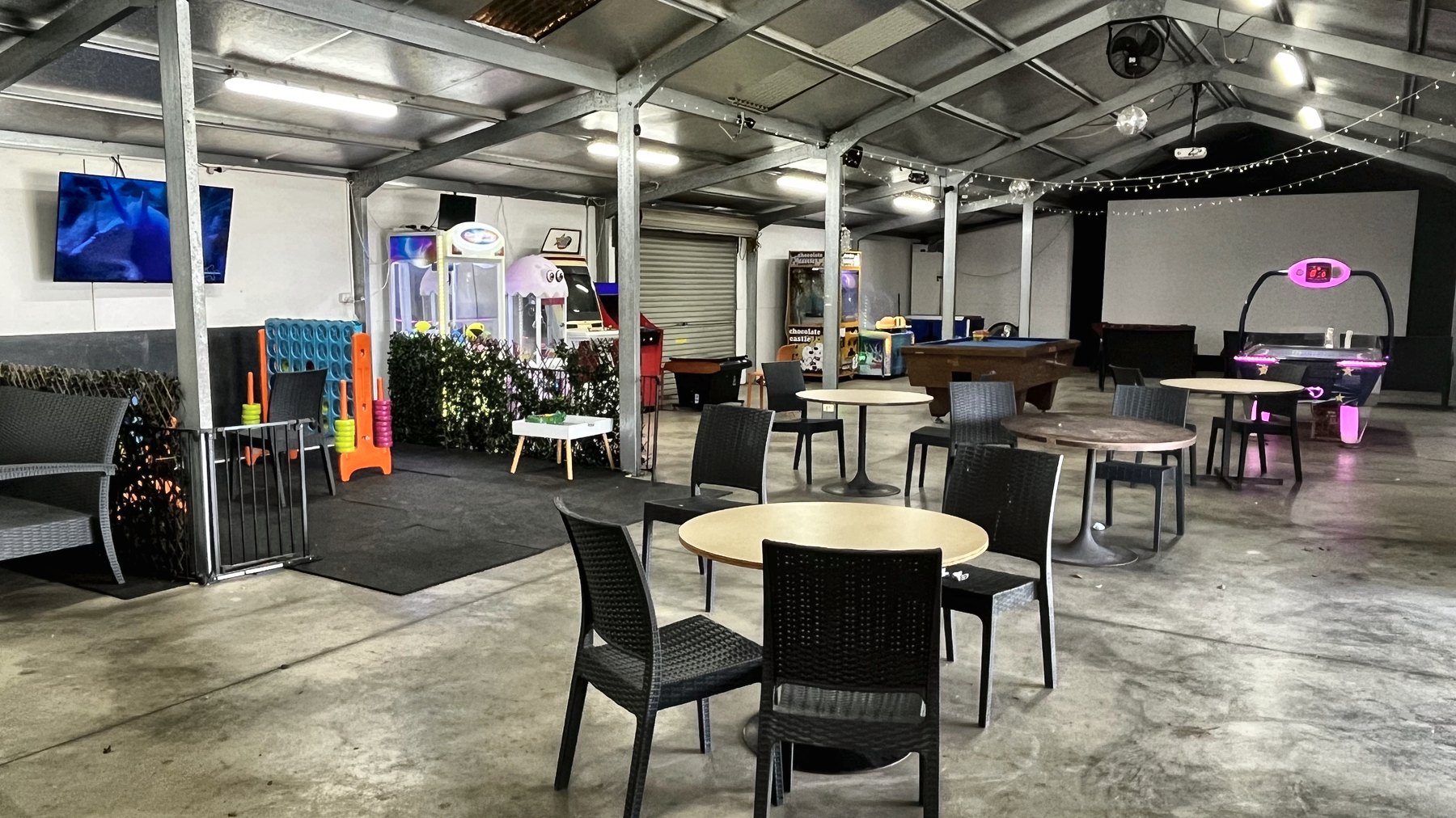 An open-sided games room with tables and chairs, a pool table, arcade machines and a wall screen