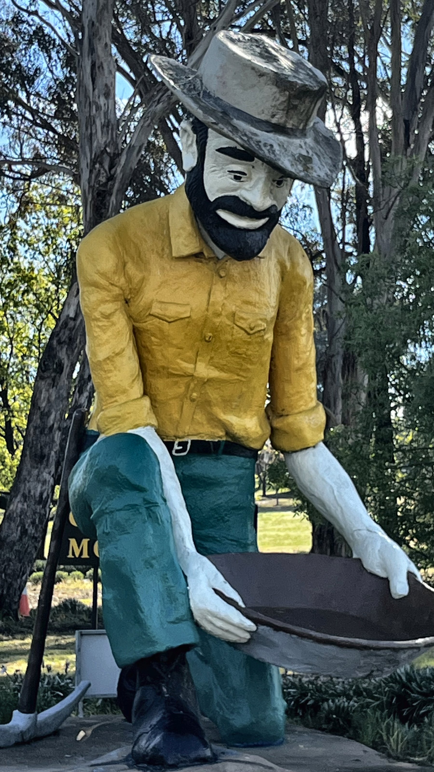 A tall painted statue of a bearded gold panner in a yellow shirt and green trousers, crouched and holding a pan