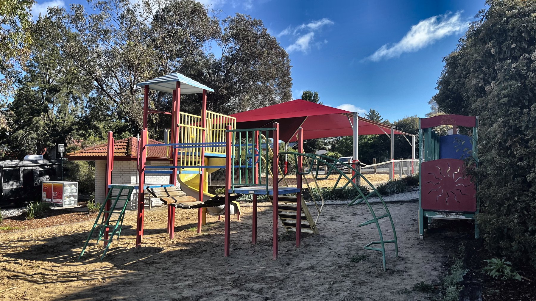 A bright steel and plastic playground frame with slides and a shade sail, on sand