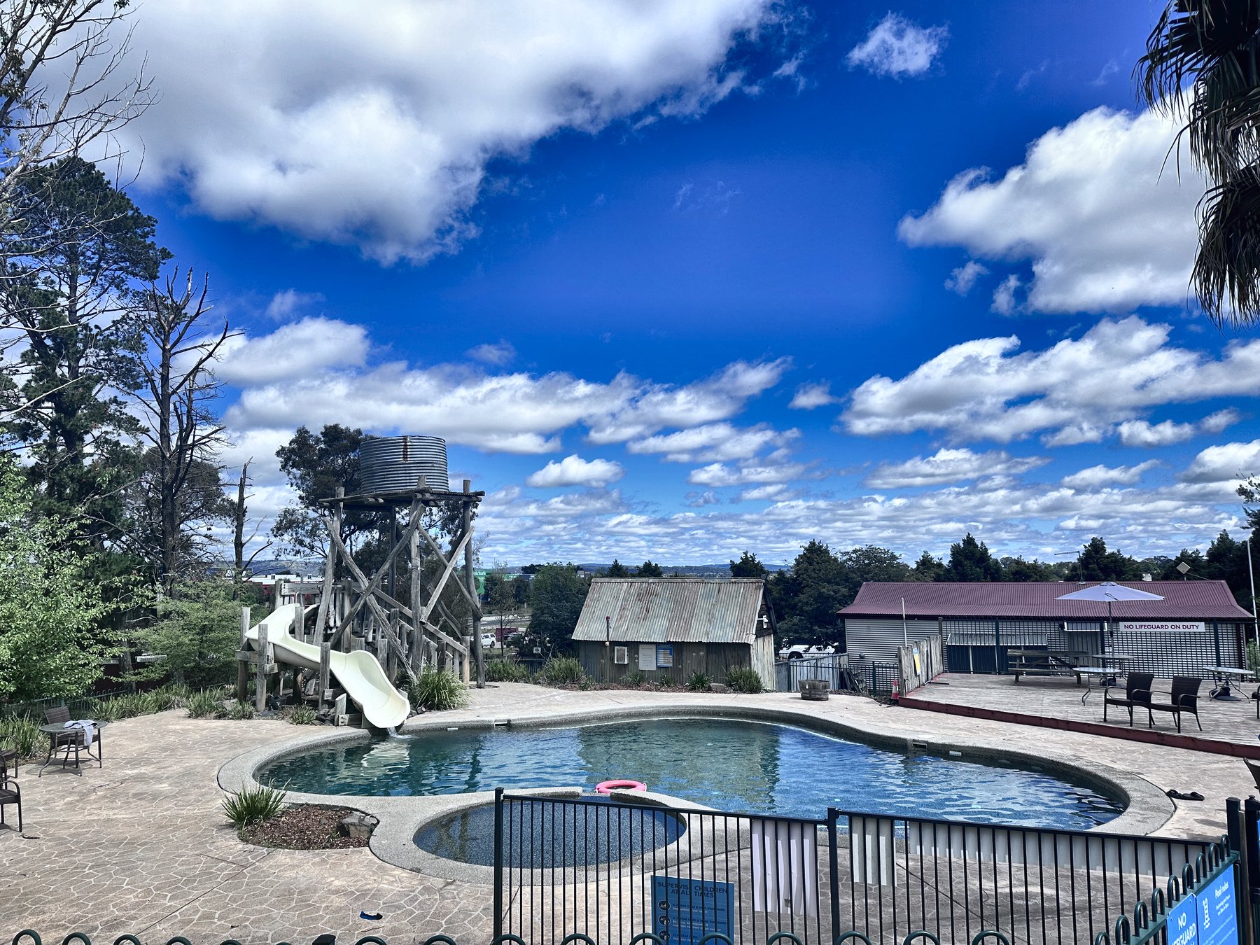 A fenced kidney-shaped swimming pool with a timber water tower and slide standing over it against a blue sky