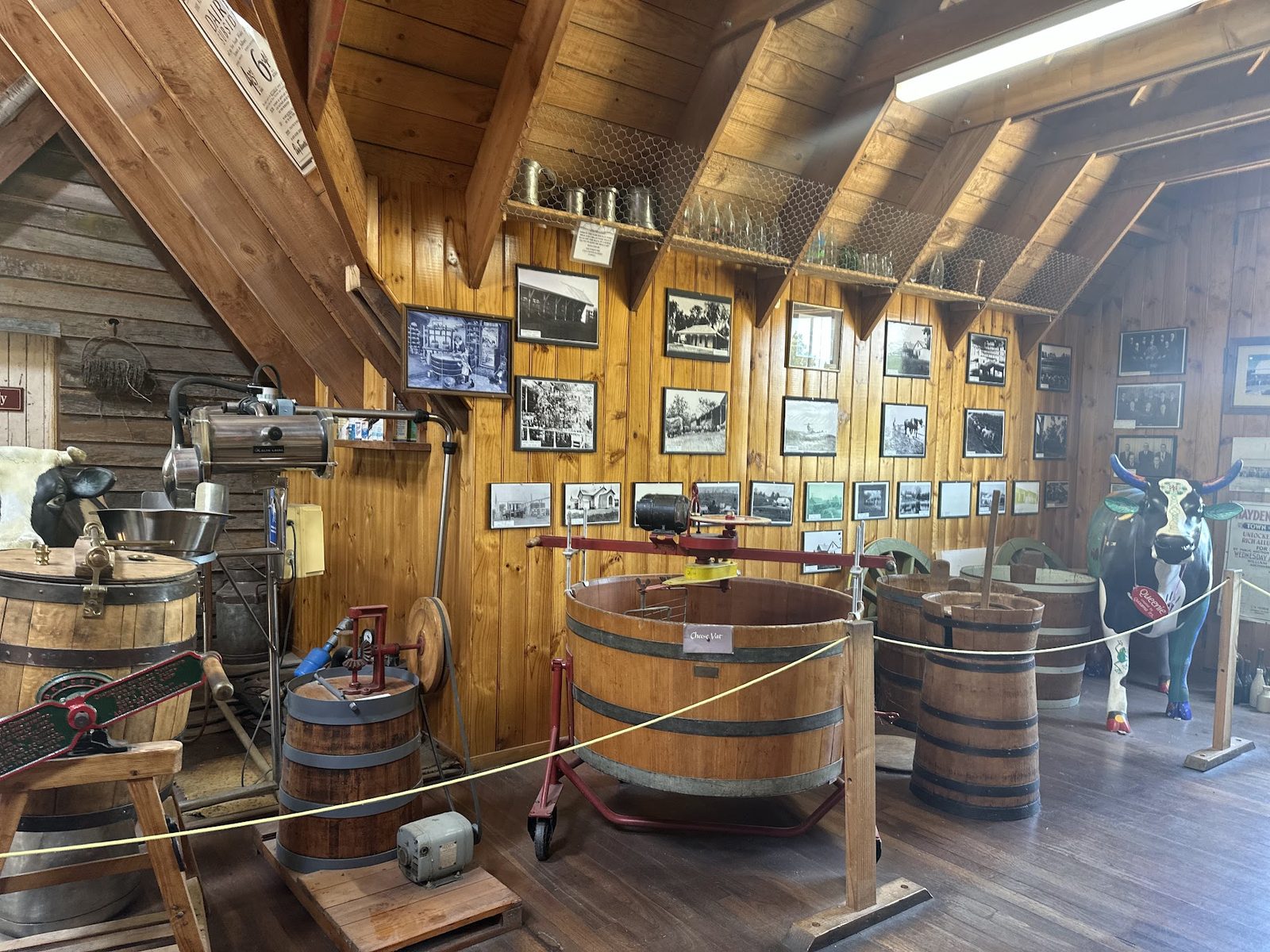 The inside of a timber slab cheese factory fitted out as a museum, with a wooden cheese vat, a cream separator, barrels and a wall of framed historic photographs