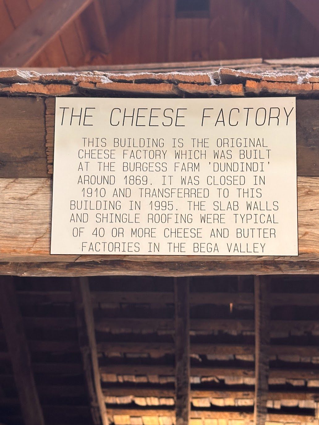 An interpretive sign fixed to a roof beam reading THE CHEESE FACTORY, explaining that the building came from the Burgess farm Dundindi