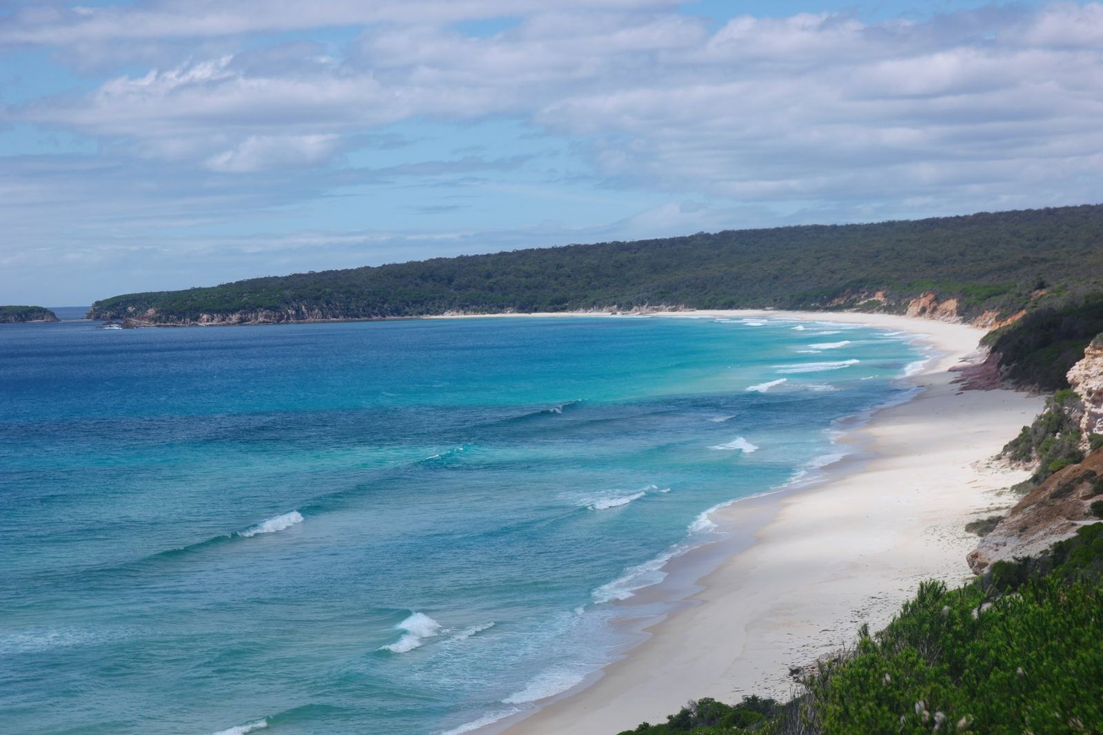 A wide bay of white sand and turquoise water backed by dense forest, an orange sandstone bluff at the near end