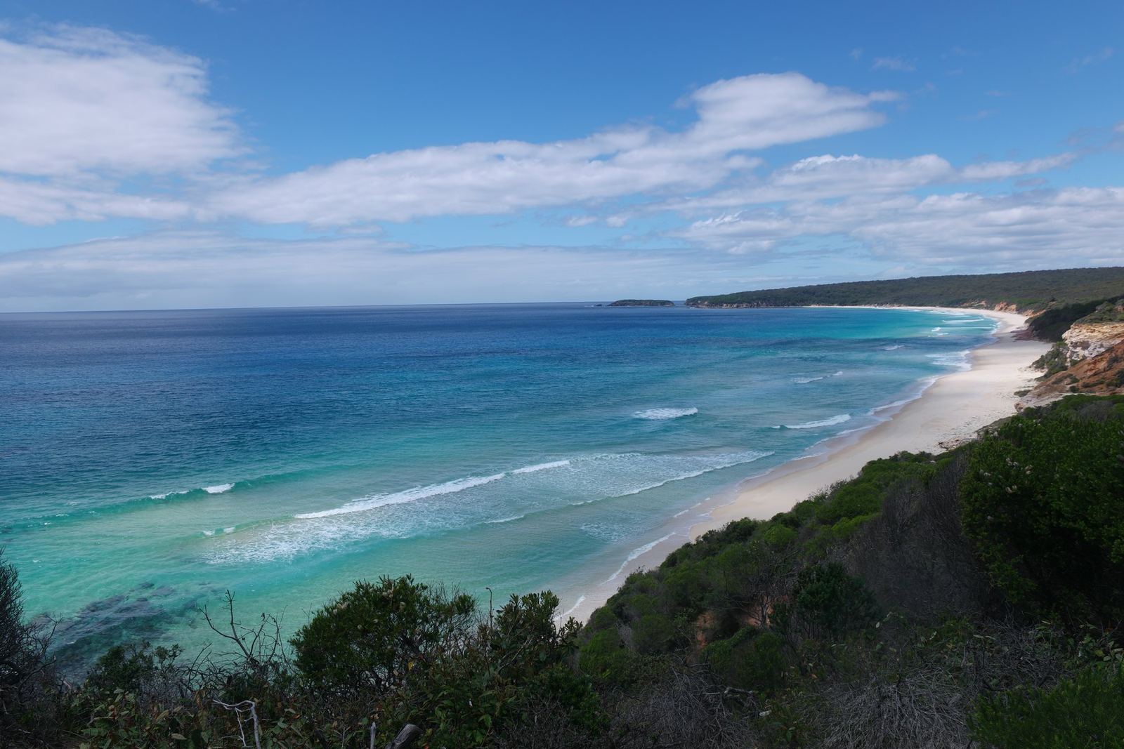 A long straight white sand beach curving away below a heath-covered clifftop, water shading from pale turquoise at the shore to deep blue offshore