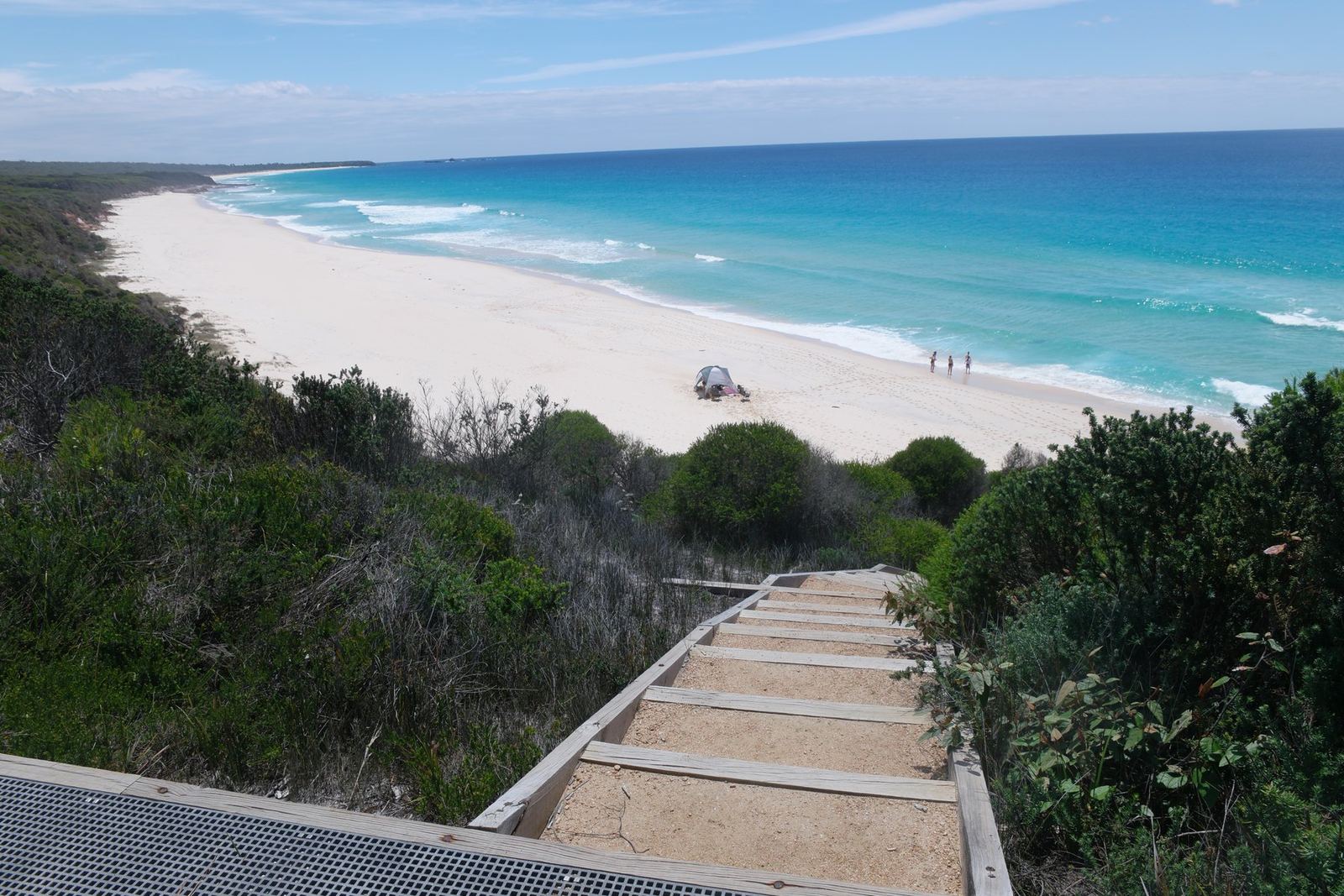 A timber boardwalk and steps descending through coastal heath towards a long white beach with two or three tiny figures on it