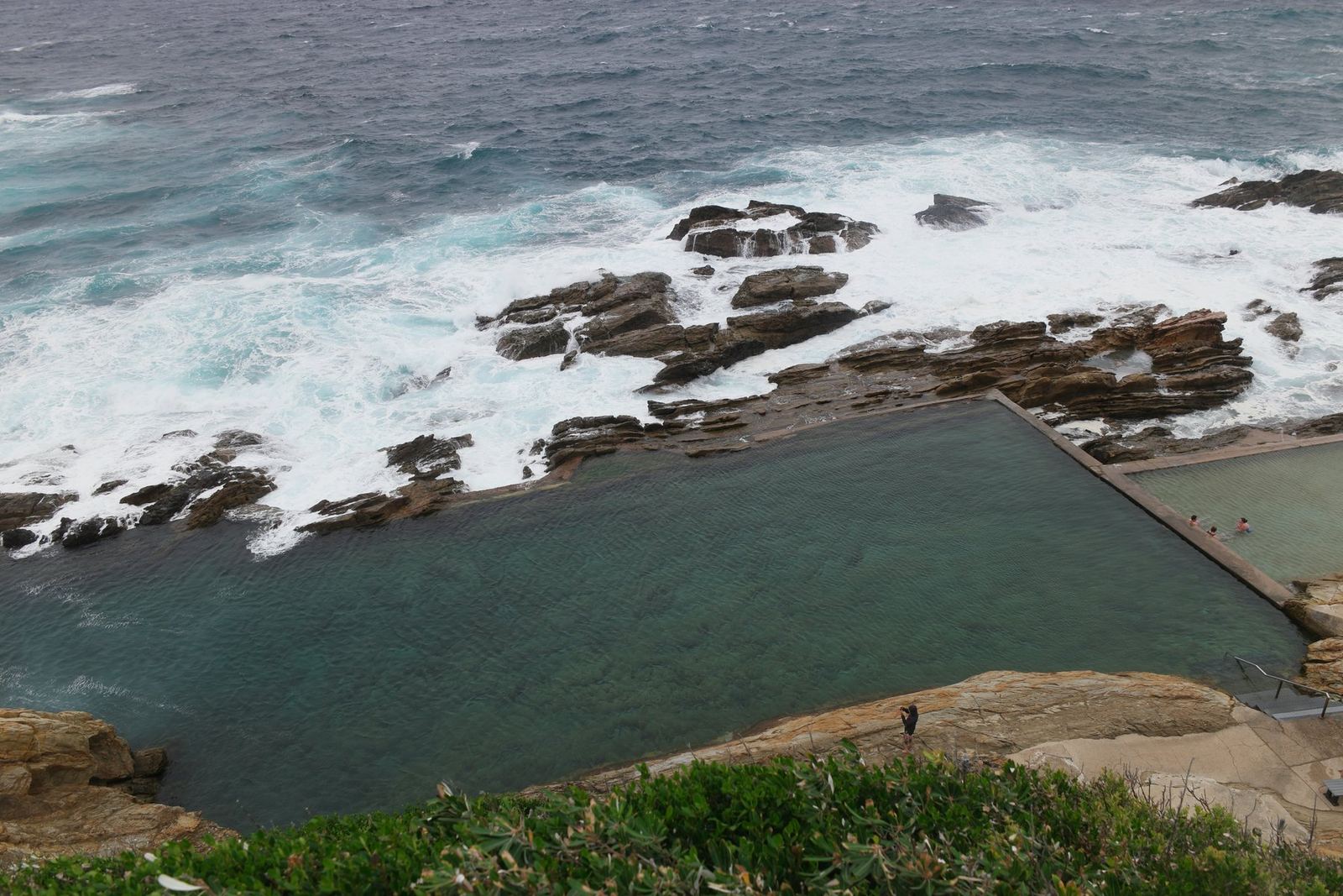 A large rectangular ocean rock pool cut into a wave-washed rock shelf below a cliff, a smaller shallow pool beside it with three swimmers, white water breaking over the rocks behind