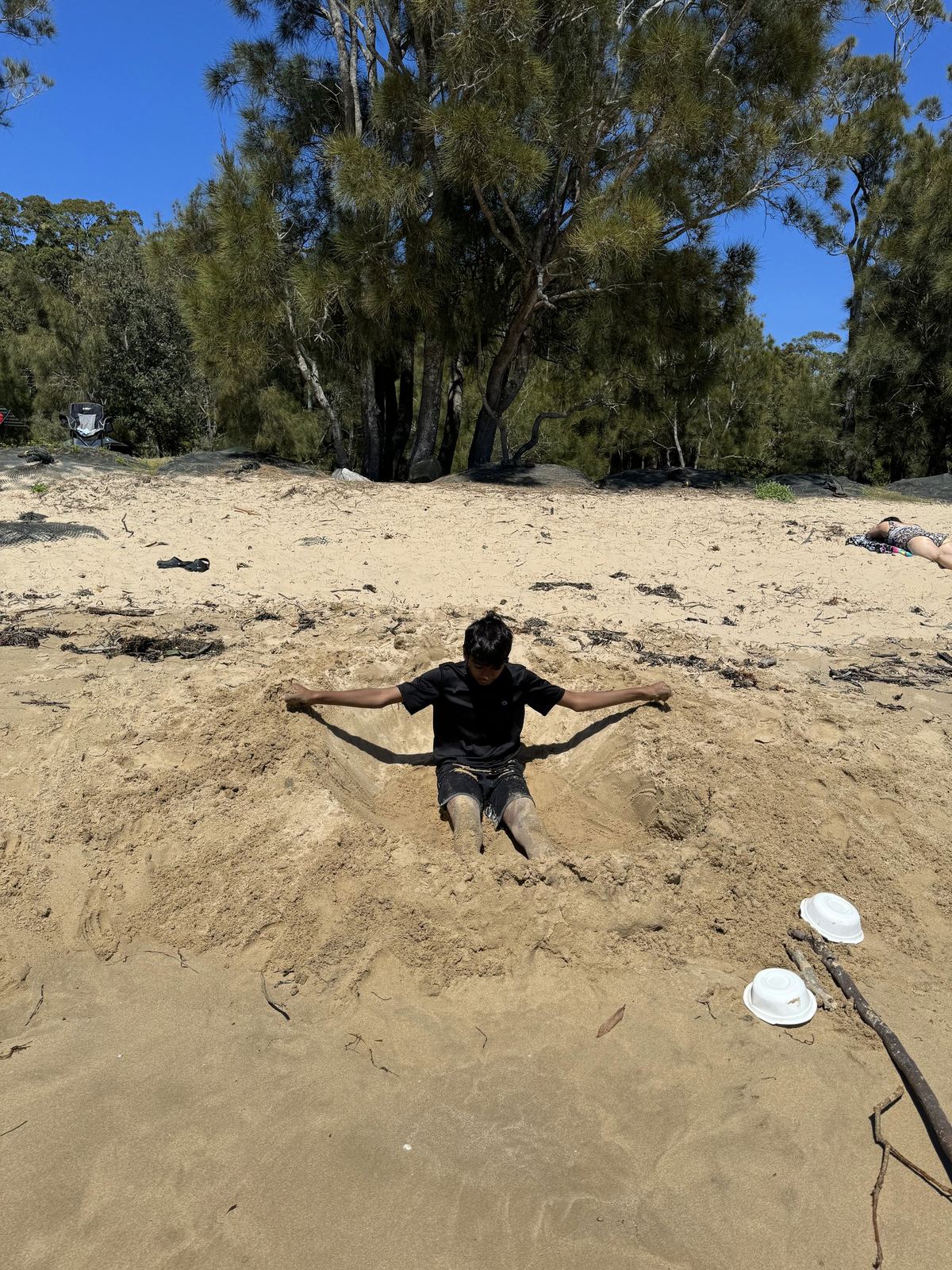 A child buried up to the chest in beach sand with arms spread wide, casuarinas and the water behind