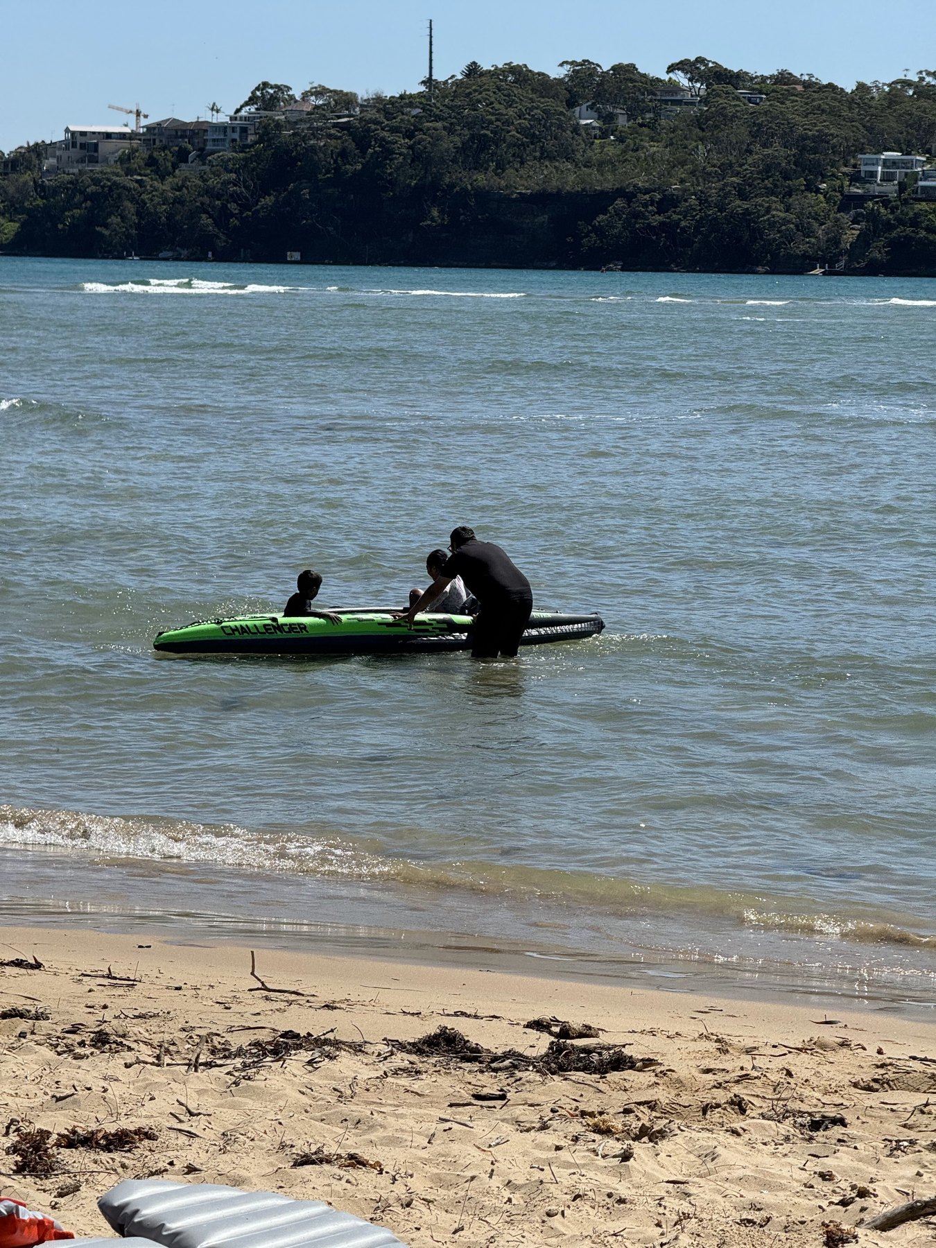 A man standing in the shallows steadying a green inflatable kayak with two children in it, the wooded far shore behind