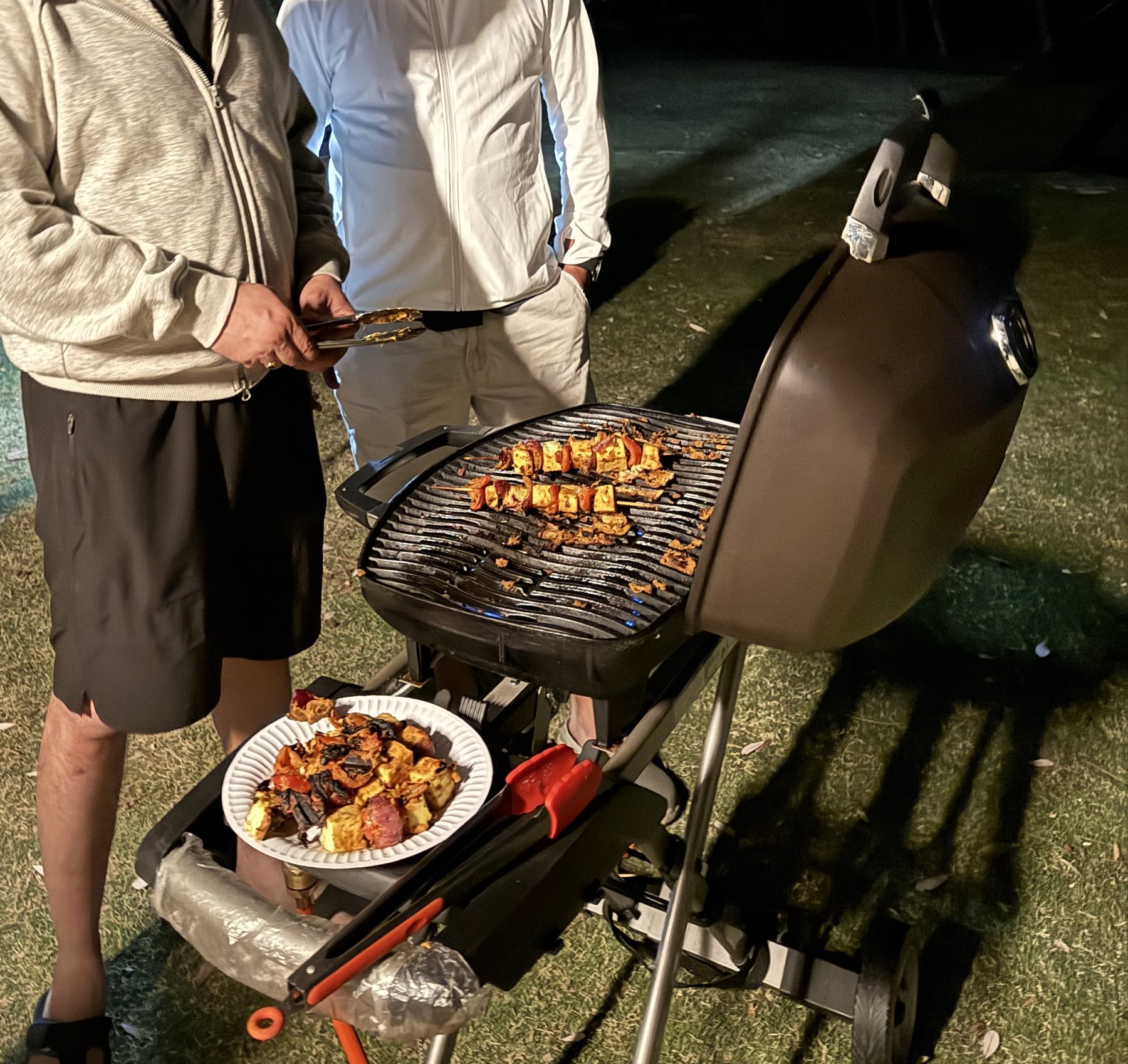 Two men seen from the chest down at a portable gas barbecue at night, paneer and vegetable skewers on the grill and a plate of them on the side table