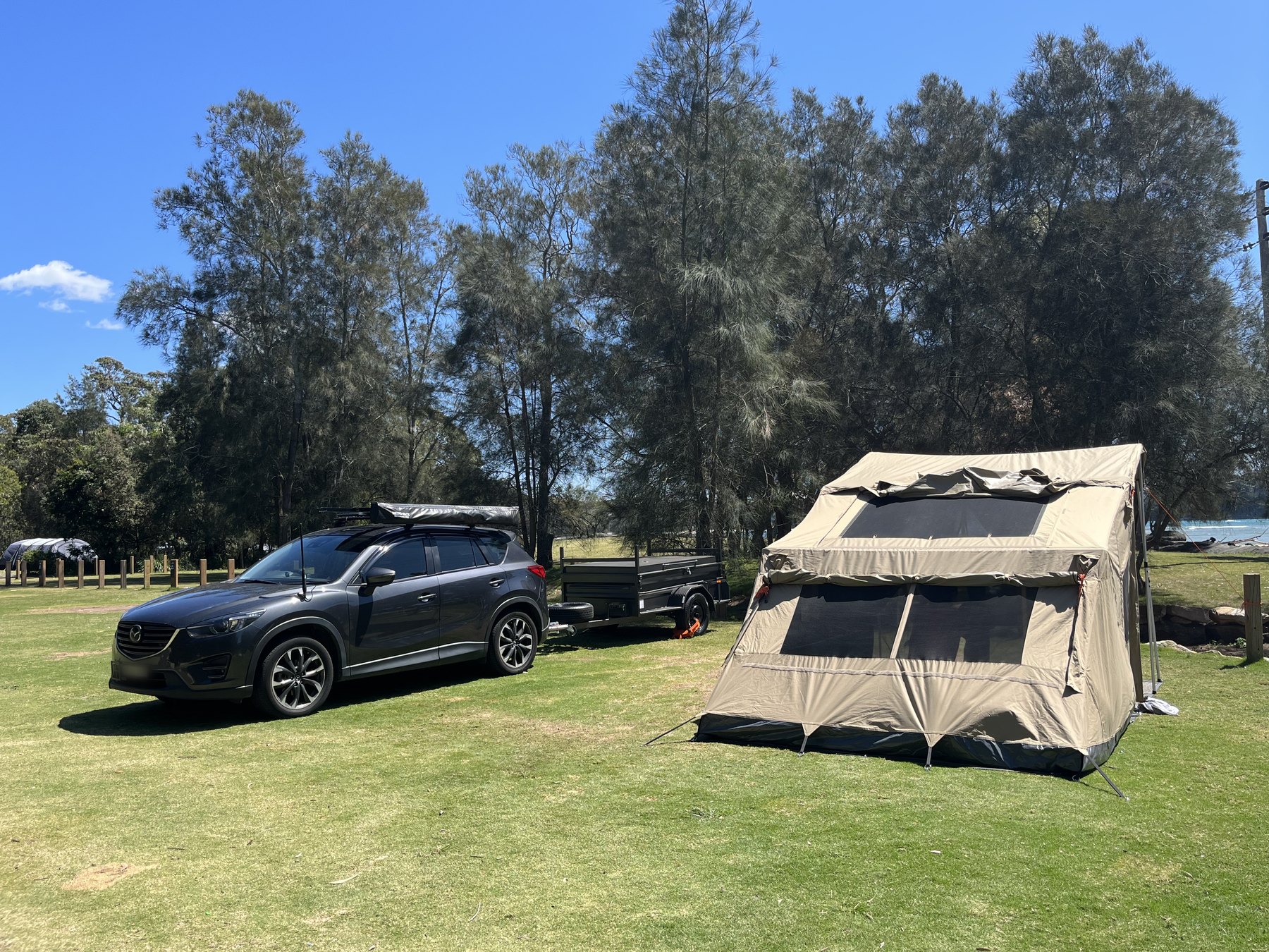 A dark SUV hitched to a box trailer beside a canvas tent on mown grass under casuarinas, the water just visible on the right