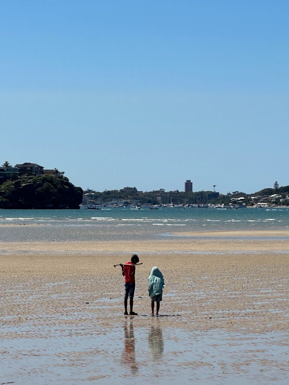 A boy standing on the wet sandflat at low tide, looking out across the bay to a wooded headland and houses on the far shore