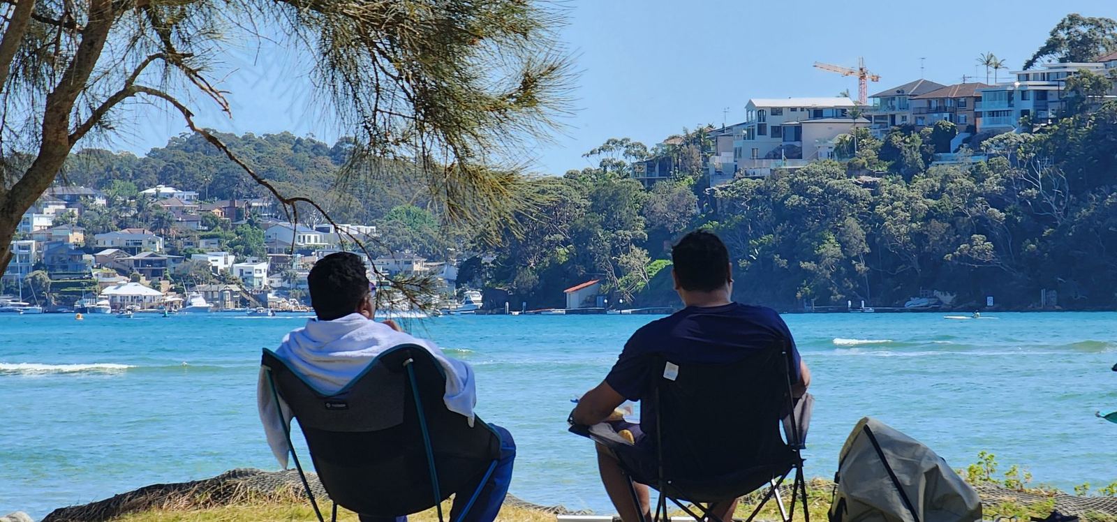Two men sitting in camp chairs at the water's edge seen from behind, looking across the bay to houses on the opposite hillside