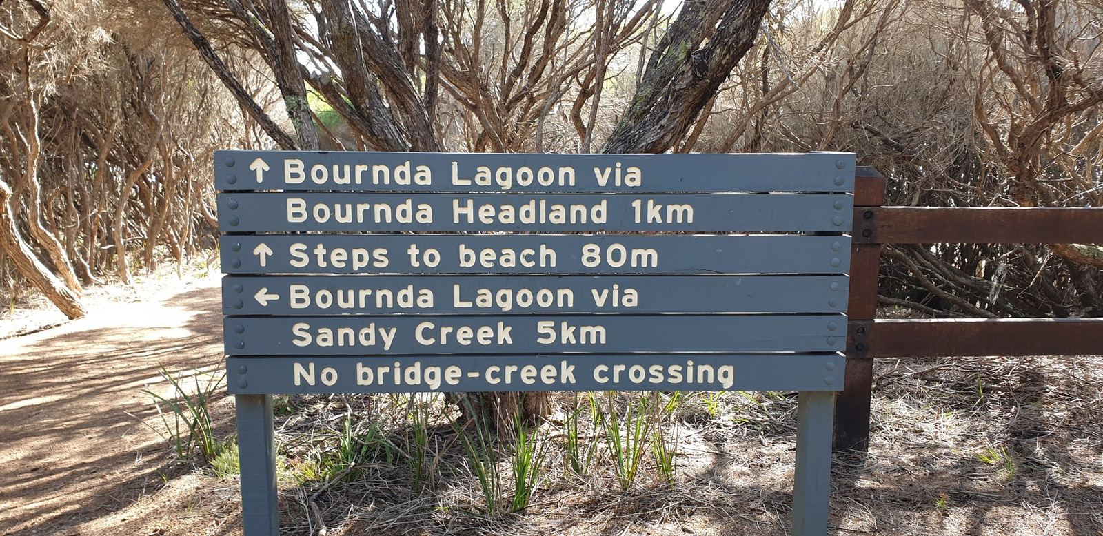 A weathered NPWS timber signpost reading Bournda Lagoon via Bournda Headland 1km, Steps to beach 80m, and Bournda Lagoon via Sandy Creek 5km no bridge creek crossing