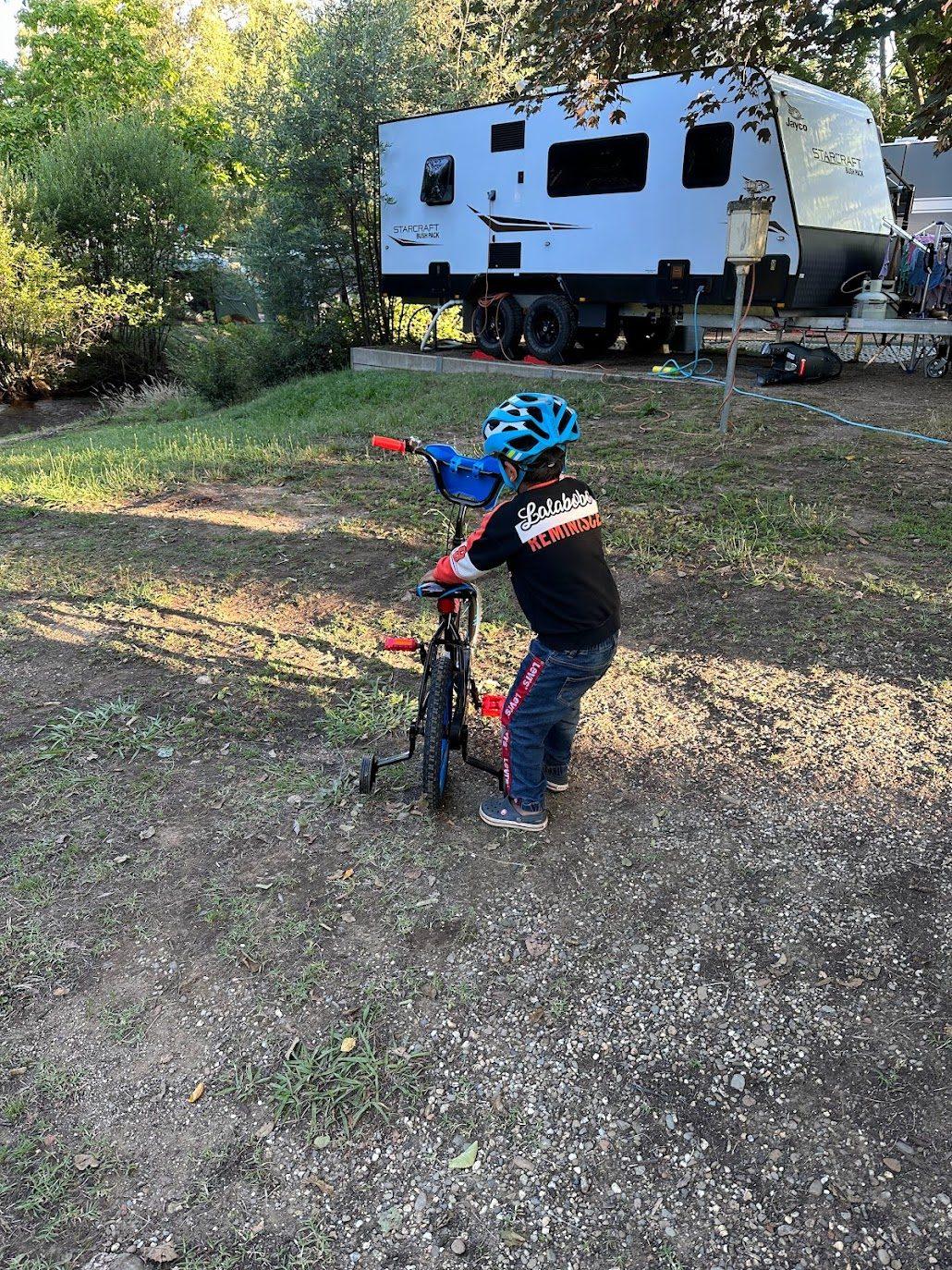 A young child riding a small bike with training wheels along the grass between caravans on a holiday park site, seen from behind
