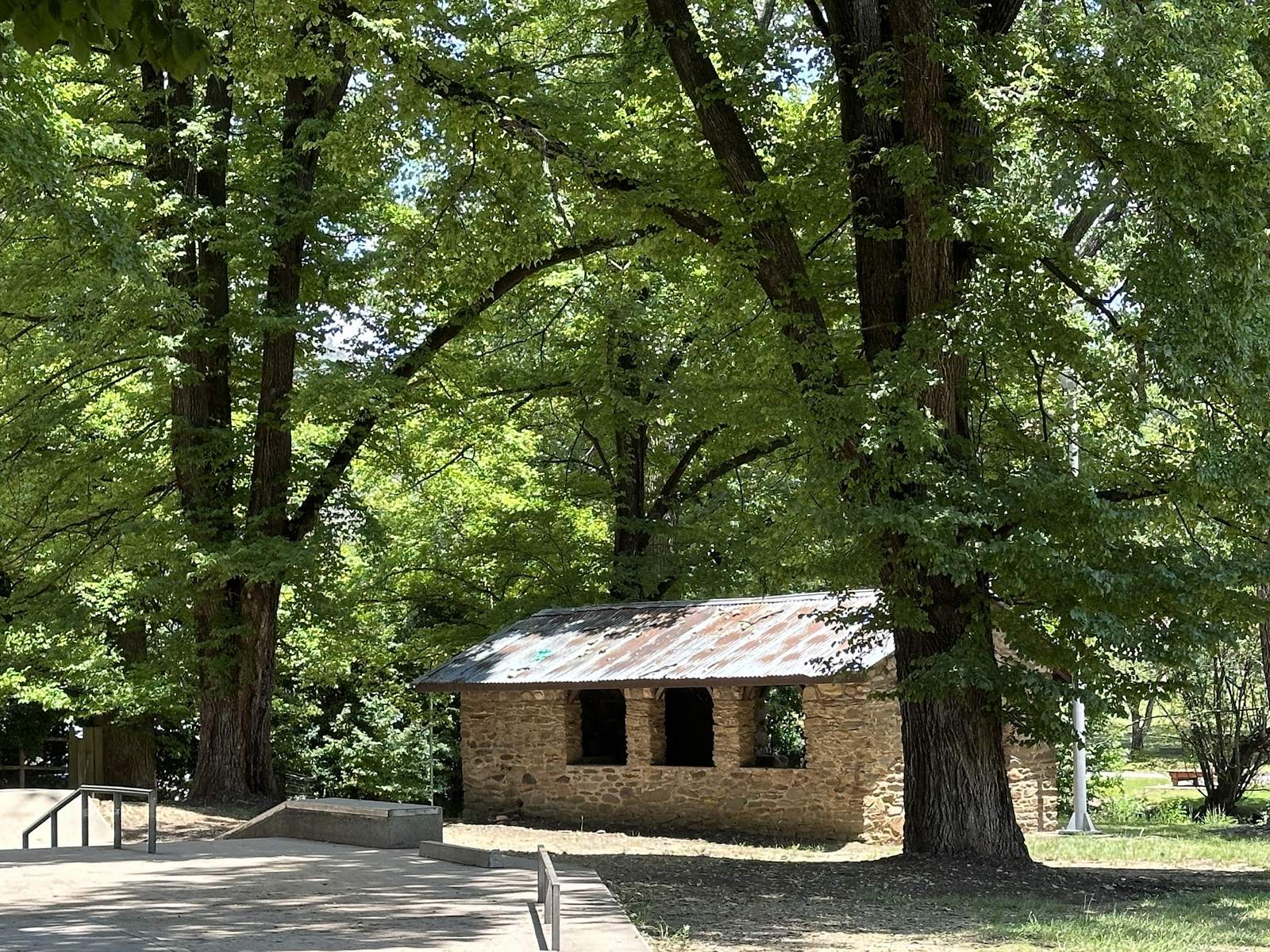 A small stone building with a rusted corrugated iron roof and open window openings, standing under tall elms beside a concrete skate ramp