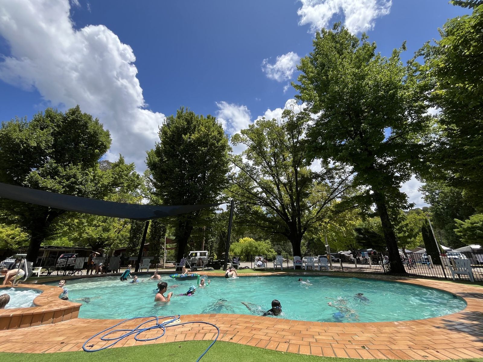 The holiday park's swimming pool under shade sails and big trees, people swimming, brick paving around the edge
