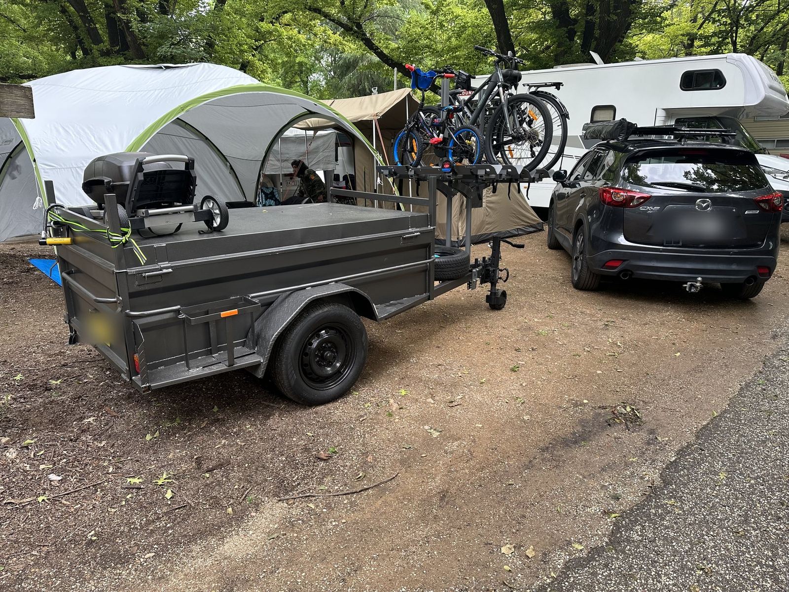 The box trailer with four bikes on its rack parked alongside the car, tents and a caravan behind on a gravel site