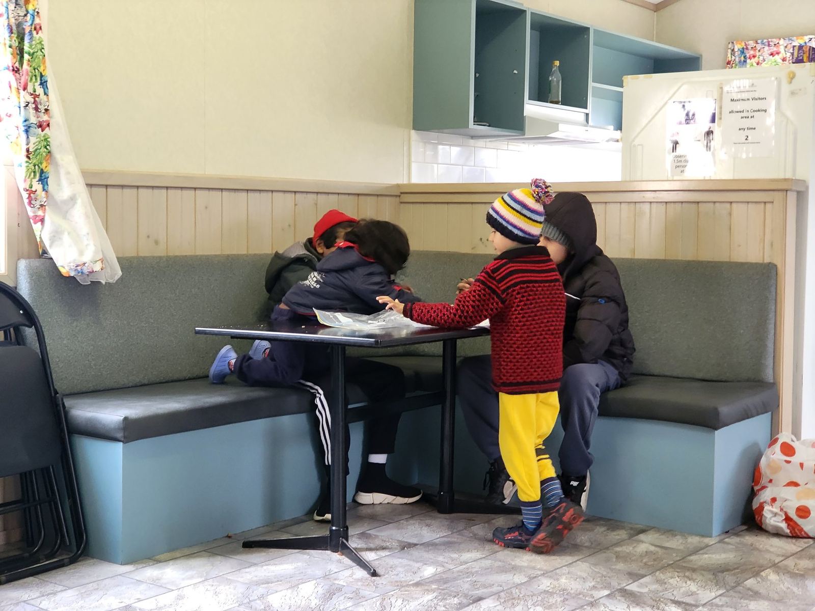Children sitting at a table in a caravan park camp kitchen, timber-lined walls, green vinyl bench seating and a kitchenette behind