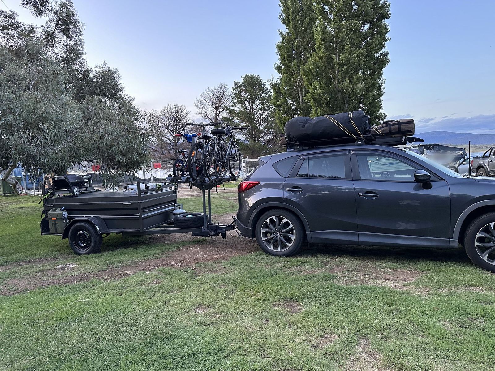 A grey SUV parked on grass with soft bags strapped to the roof rack, towing a high-side box trailer carrying three bikes on a rack and a jerry can