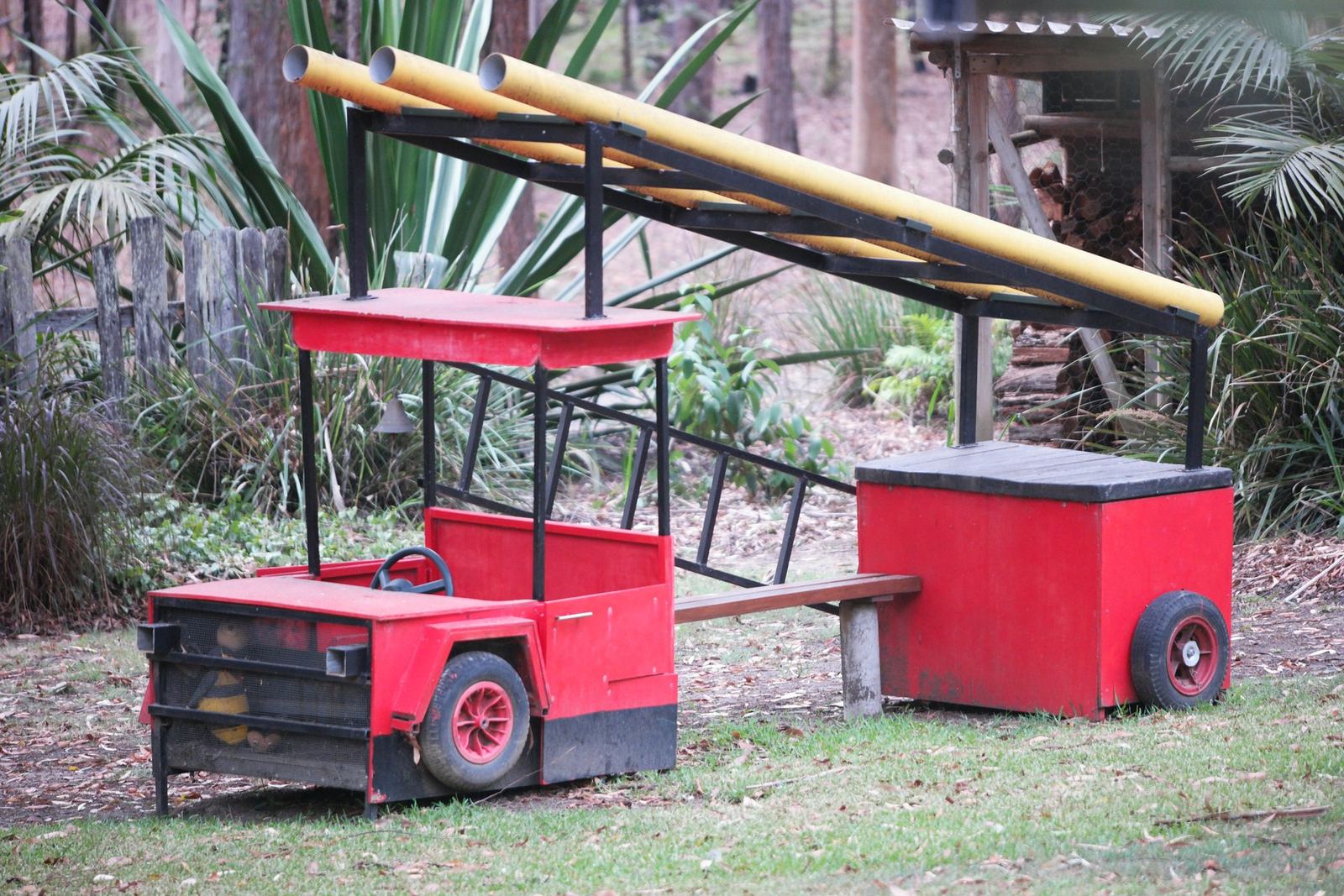 Two red pedal karts and a red bin standing on the grass in front of a long yellow-railed ramp