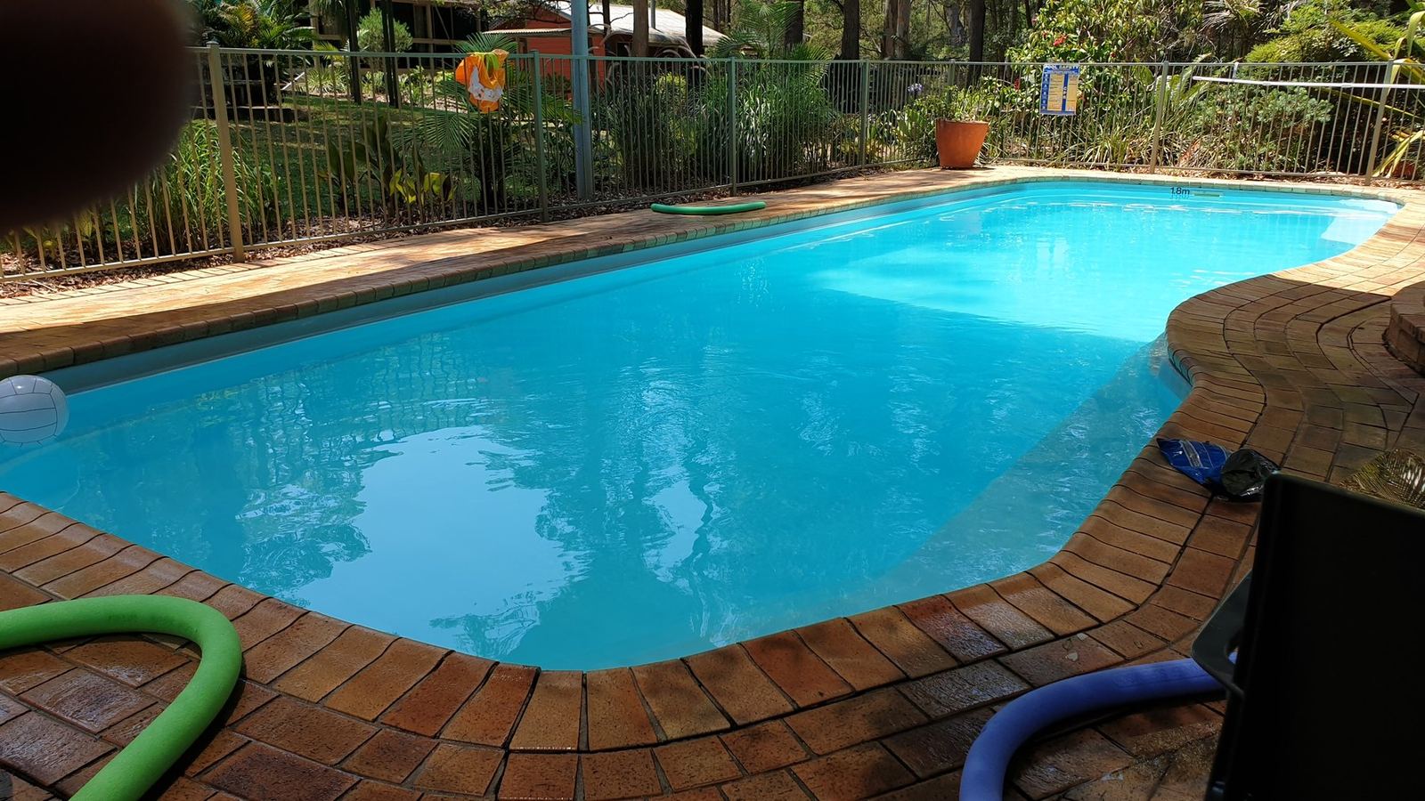 The pool from the deck, bright blue water inside a brick surround with a pool hose coiled at the edge