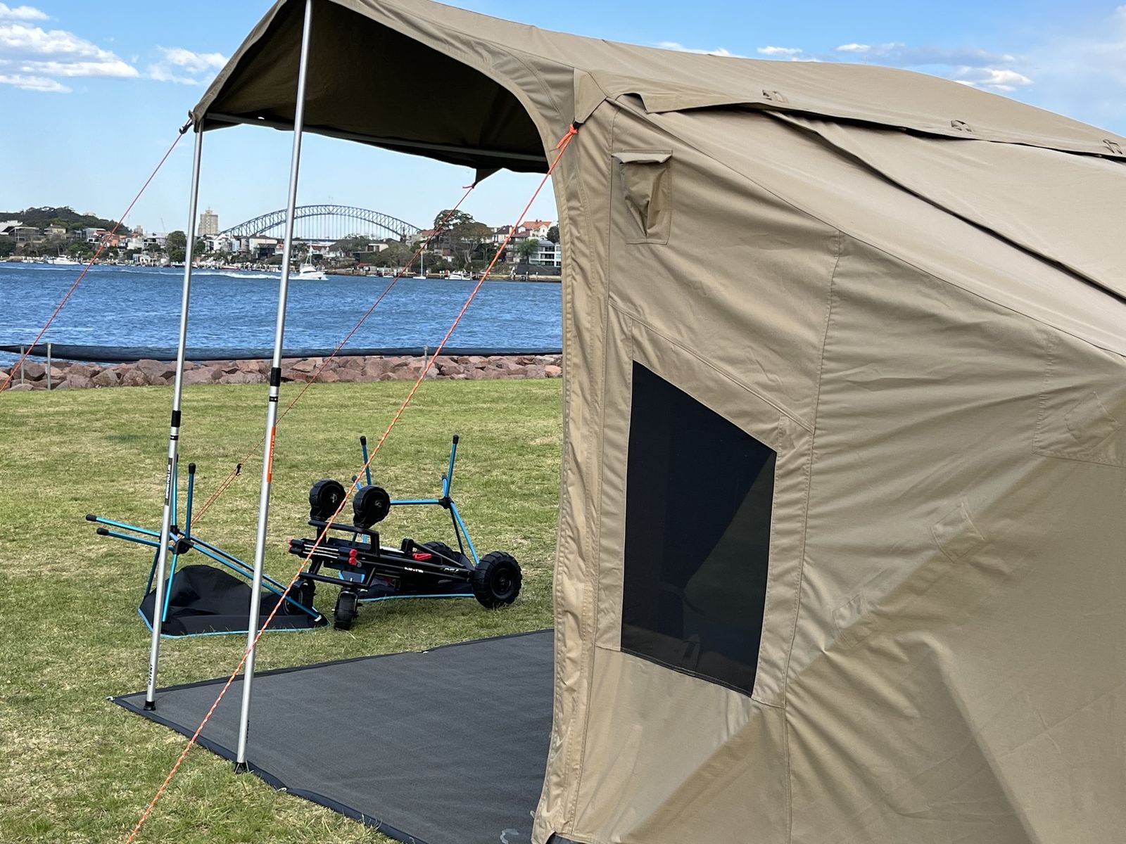 A canvas tent with its awning up on mown grass at the water's edge, the Sydney Harbour Bridge on the skyline behind it