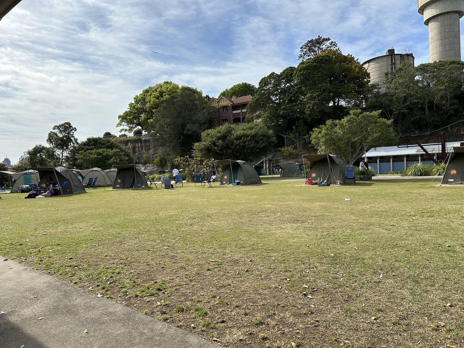 The same row of safari tents seen from further along the grass, with the cut sandstone cliff and a house on the ridge above