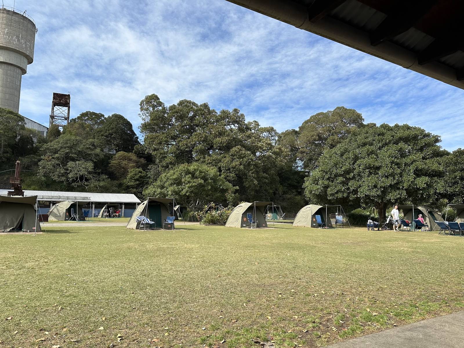 A row of canvas safari tents pitched on mown grass beneath a large fig, with a steel water tower and a crane behind them