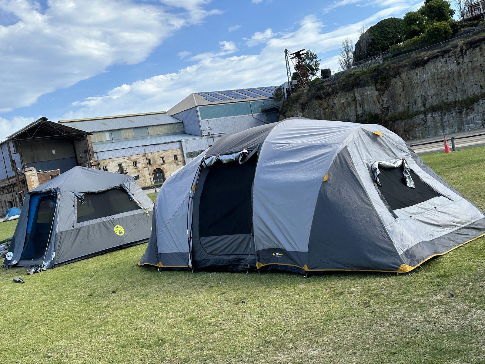 Two dome tents on grass in front of a long industrial shed with solar panels, the sandstone cliff and a crane rising behind
