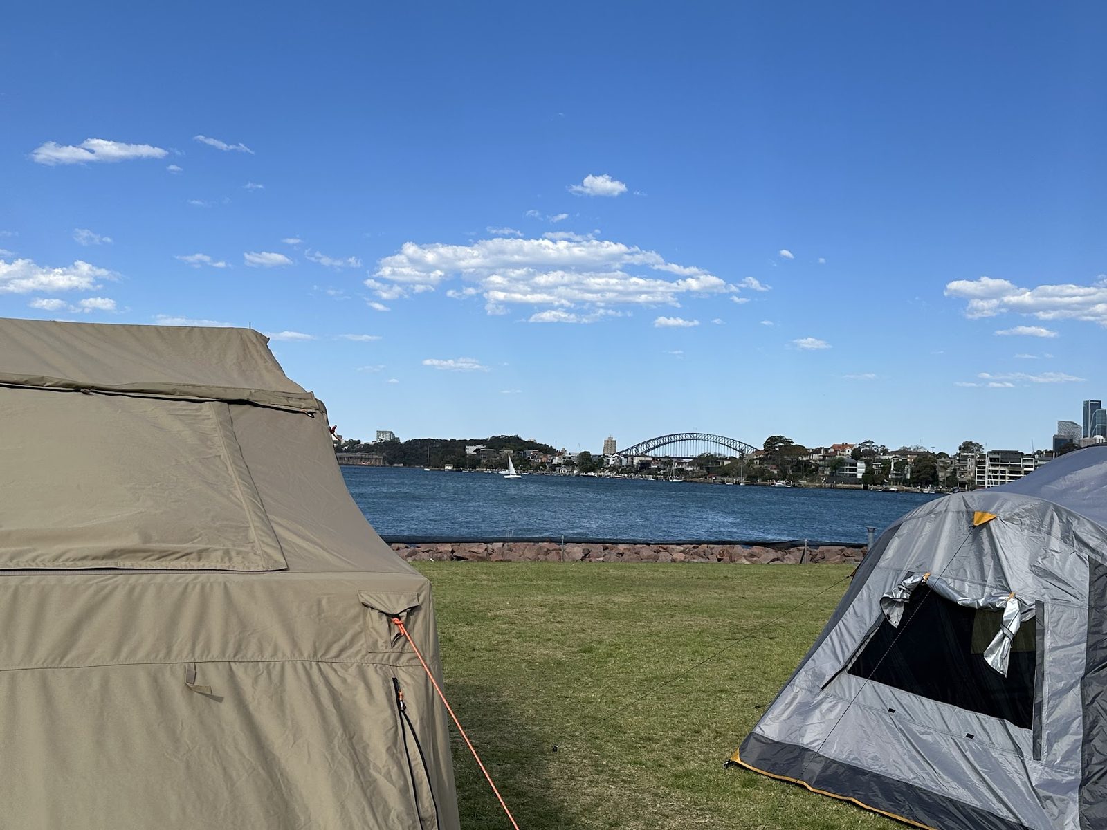 Two tents on the grass by the water with a yacht passing and the Sydney Harbour Bridge small on the horizon behind