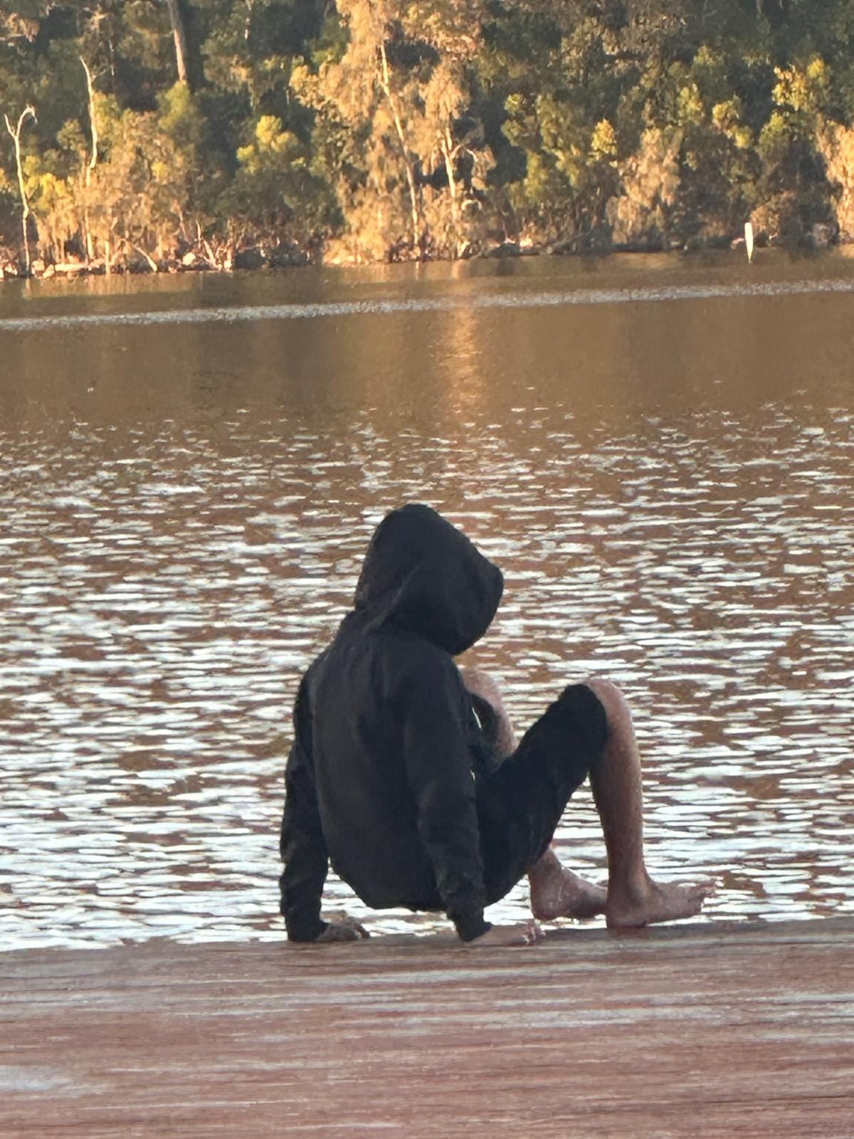 Someone in a black hooded jacket sitting on the concrete boat ramp at the edge of the water, back to the camera