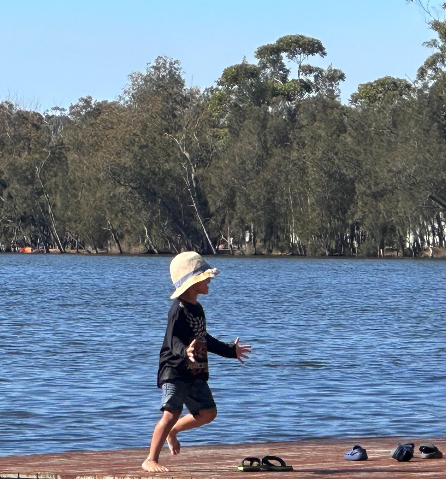 A child in a wide-brimmed hat walking into the shallows at the edge of the lake, thongs left on the sand behind