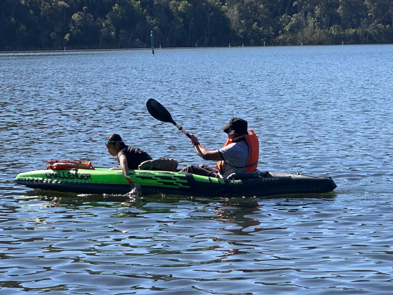 Two people paddling a green inflatable kayak on flat water, the far shore wooded behind them