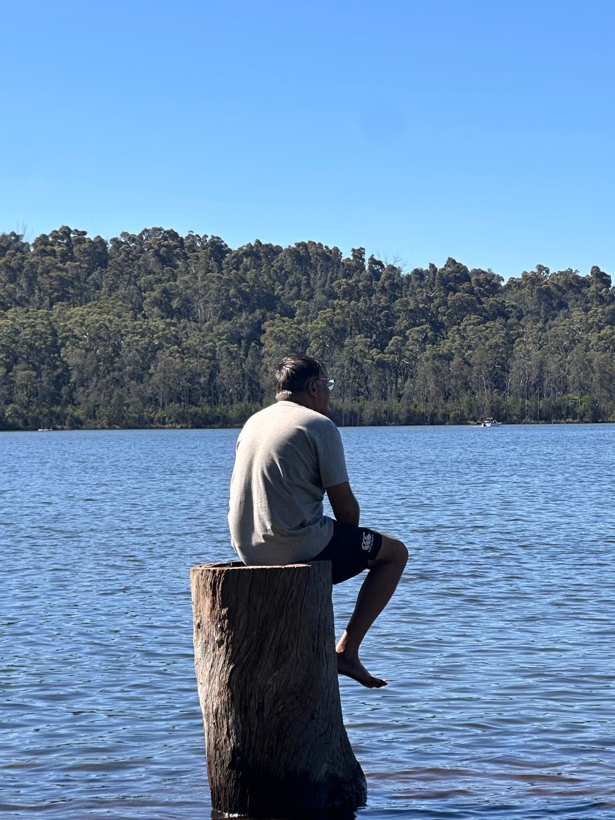 A man sitting on top of a weathered timber mooring post at the water's edge, seen from behind, looking across the lake to a wooded ridge