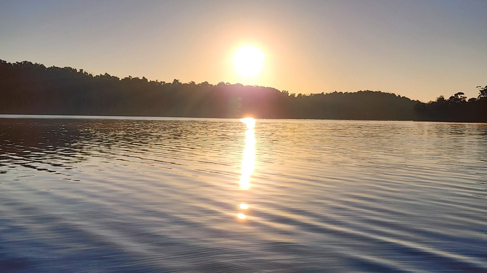Sunrise over Lake Conjola, the sun low over a wooded far shore and a long column of light down the still water