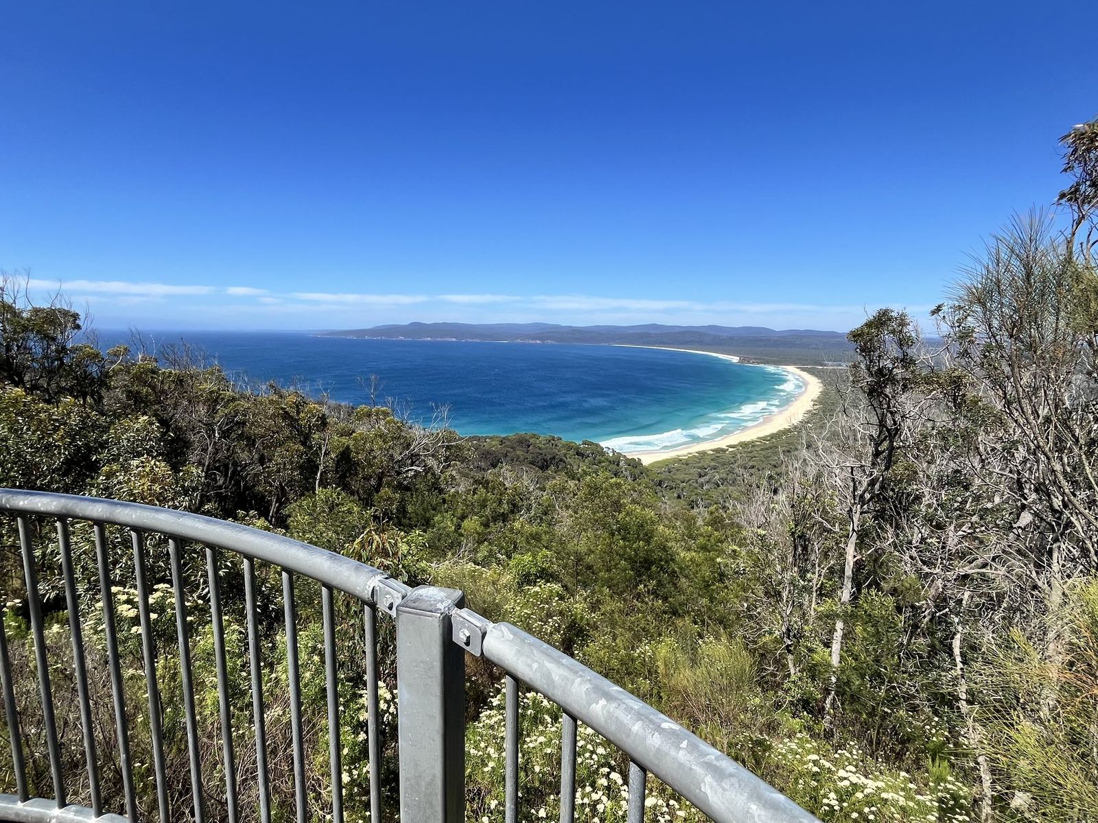 The same bay framed by scrub and a lookout railing, the beach a thin white line in the distance