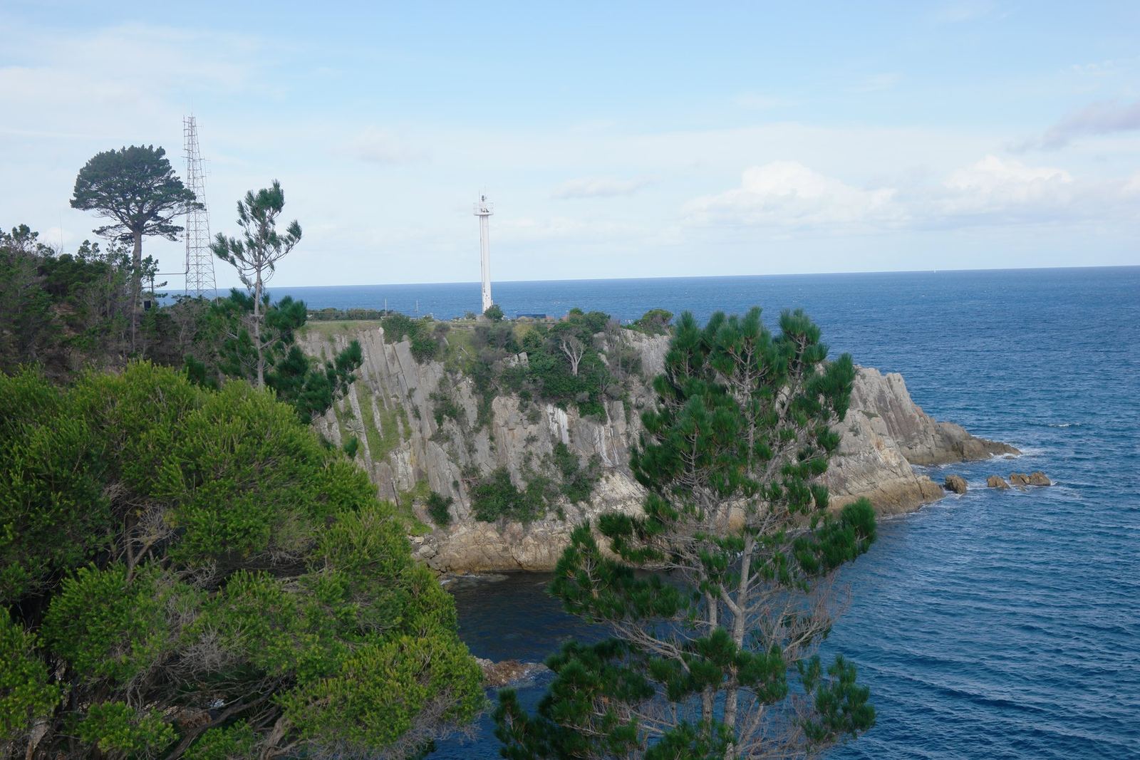 A white lattice navigation light tower standing on a grey rocky headland above the sea, dark pines in the foreground and deep blue ocean beyond