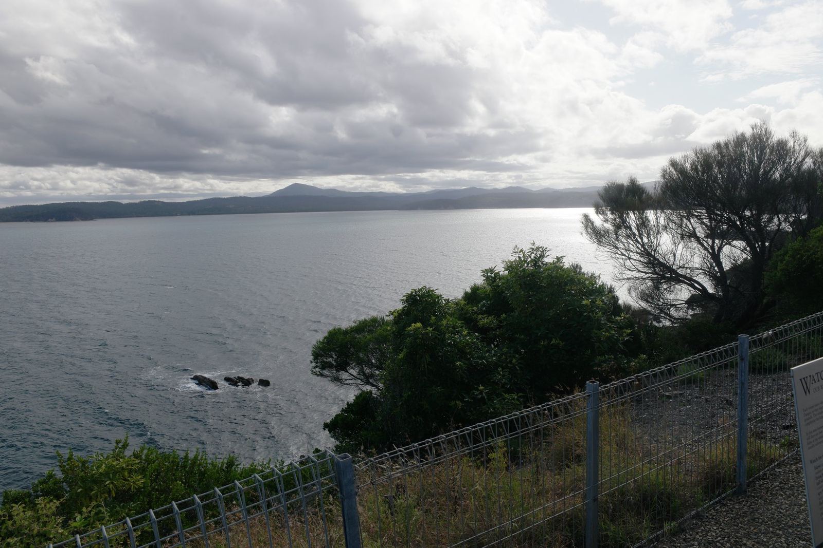 The same bay from behind a mesh safety fence, with the edge of a whale-watching interpretive sign in the frame