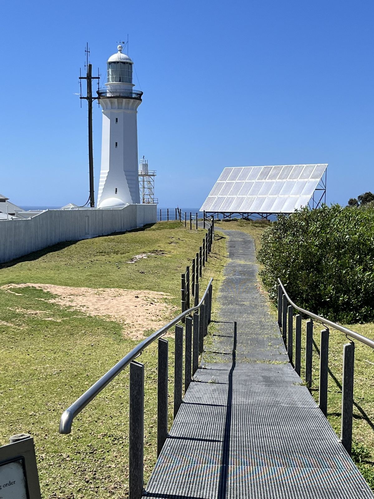A boardwalk running towards Green Cape lighthouse across open headland grass, solar panels beside the path